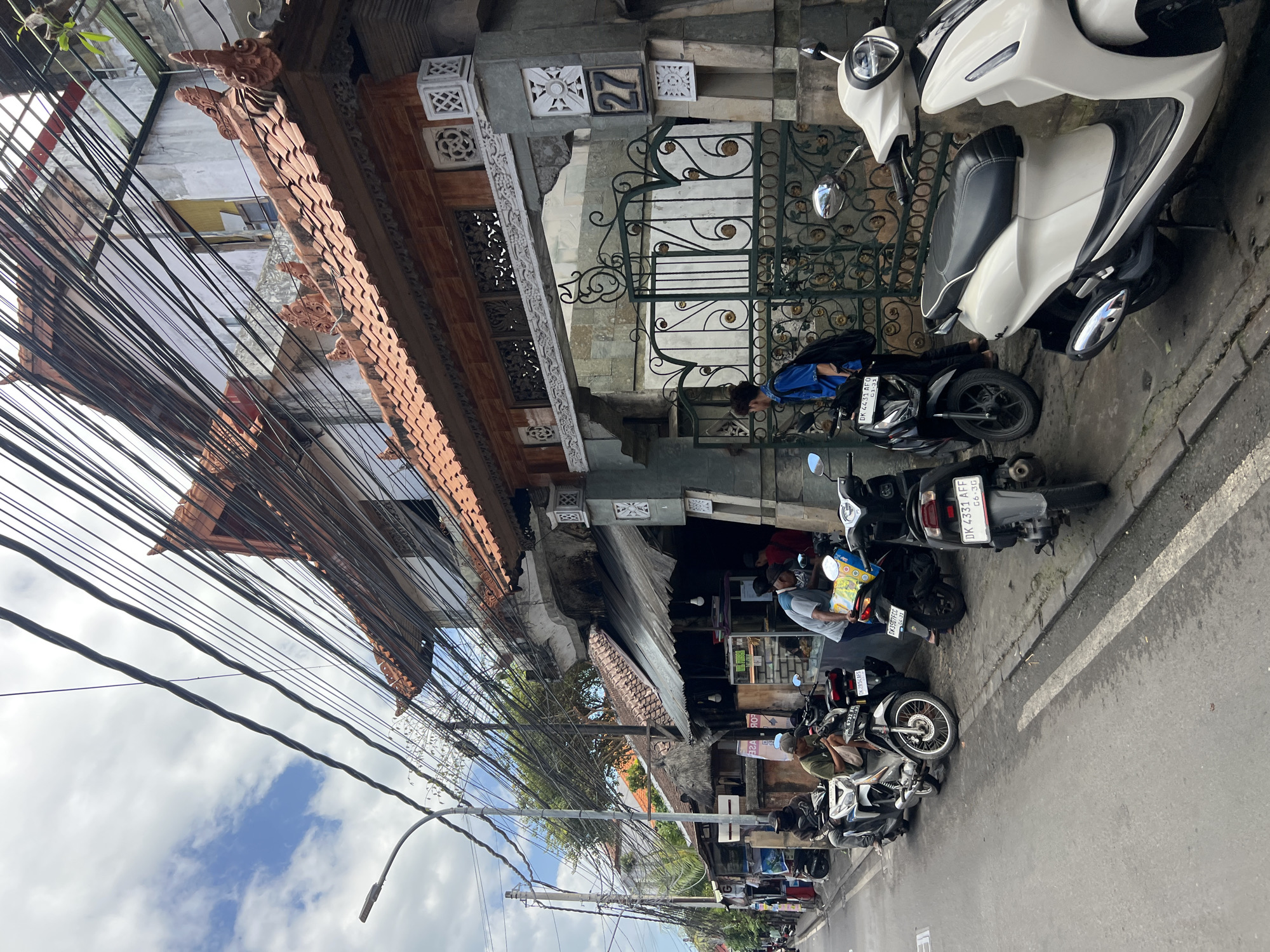 A street scene in Kuta, Indonesia, shows a row of parked motorcycles and scooters along a narrow sidewalk. A building with a traditional tiled roof and intricate carvings stands on the right, displaying the number "27" on its facade. A green wrought-iron gate, adorned with decorative scrollwork, is partially visible. Several individuals are interacting with the motorcycles and a small storefront. The street is paved, and a lamppost is present on the left. Overhead, a dense network of electrical wires crisscrosses the sky, obscuring the buildings and sky. The overall impression is a bustling urban environment with elements of local architecture and everyday life.