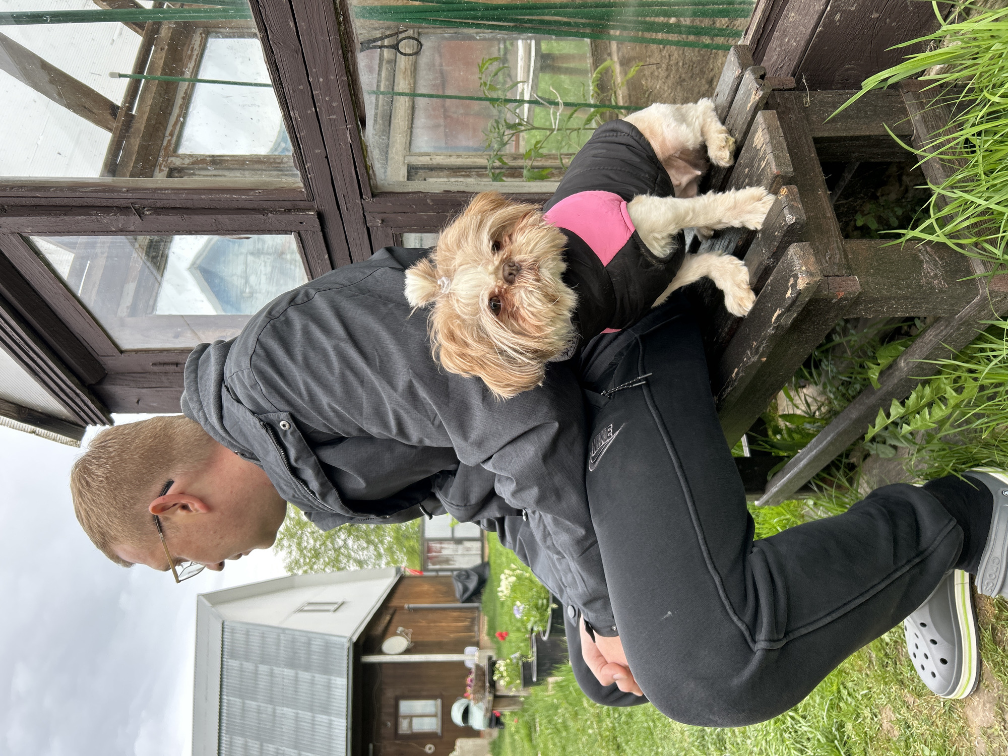 A man kneels outdoors near a wooden structure, with a small dog perched on his back. The man wears a gray jacket, black sweatpants with a white Nike logo, black socks, and gray Crocs. He has short, blond hair and wears glasses. The dog is small and light-colored with long fur, wearing a black and pink jacket. They are next to a weathered wooden structure that appears to be part of a greenhouse or small building. The background shows a green grassy area, other buildings, and overcast skies. Based on associated geotags and other data, this scene is likely located in Meshchovsk, Russia.