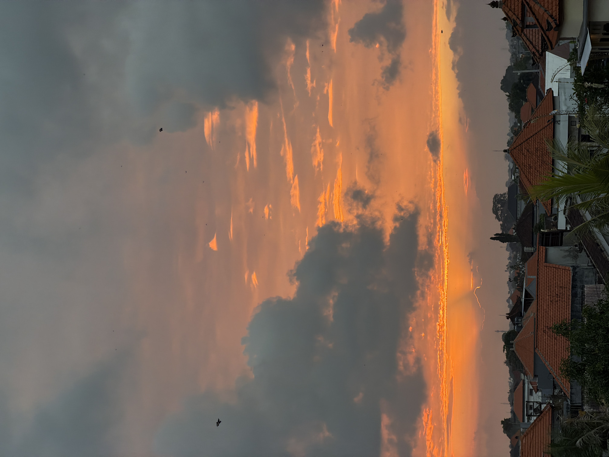 The image depicts a sunset scene over the city of North Kuta, Indonesia. The sky is filled with dramatic clouds, with hues of orange and pink from the setting sun creating a vibrant contrast with the darker, gray clouds. The sun's rays are visible breaking through the clouds near the horizon. Below the sky, a cityscape is visible, characterized by numerous buildings with distinctive orange-tiled roofs, typical of Indonesian architecture. Some of the buildings appear to be residential houses, while others might be commercial or public structures. Palm trees and other foliage can be seen interspersed among the buildings, adding a touch of tropical greenery. A few birds are visible as small dark specks in the sky, and one bird is captured in flight in the upper left quadrant of the image. The overall impression is that of a serene, late afternoon or early evening moment in a bustling urban environment with natural beauty.