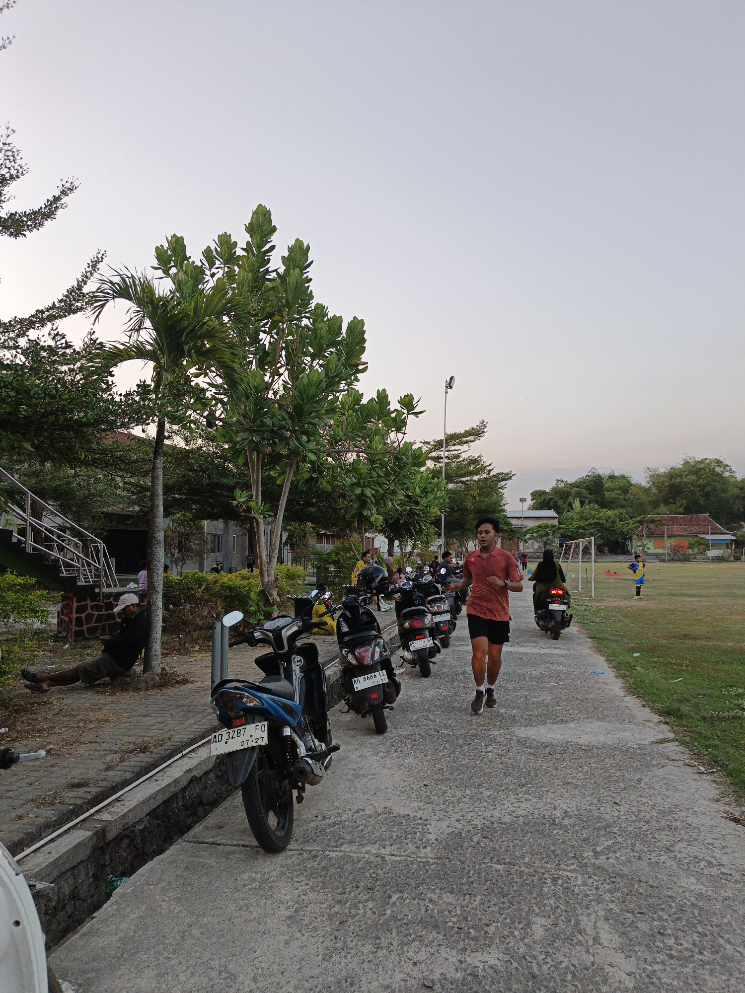 The image depicts an outdoor scene in Planggu, Indonesia, likely in the late afternoon or early evening given the lighting. In the foreground, a young man in a red t-shirt and black shorts is jogging on a paved path. Behind him, several motorcycles are parked along the left side of the path. Further back, on the left, a person is sitting against a tree, leaning on a low wall. A set of stairs with a metal railing is visible behind the sitting person. On the right side of the path, a grassy field stretches out, with a soccer goal visible in the distance. A few other individuals are present on the field and near the parked motorcycles. The background consists of trees, some buildings, and a pale sky. The overall atmosphere suggests a community space used for recreation and daily activities.