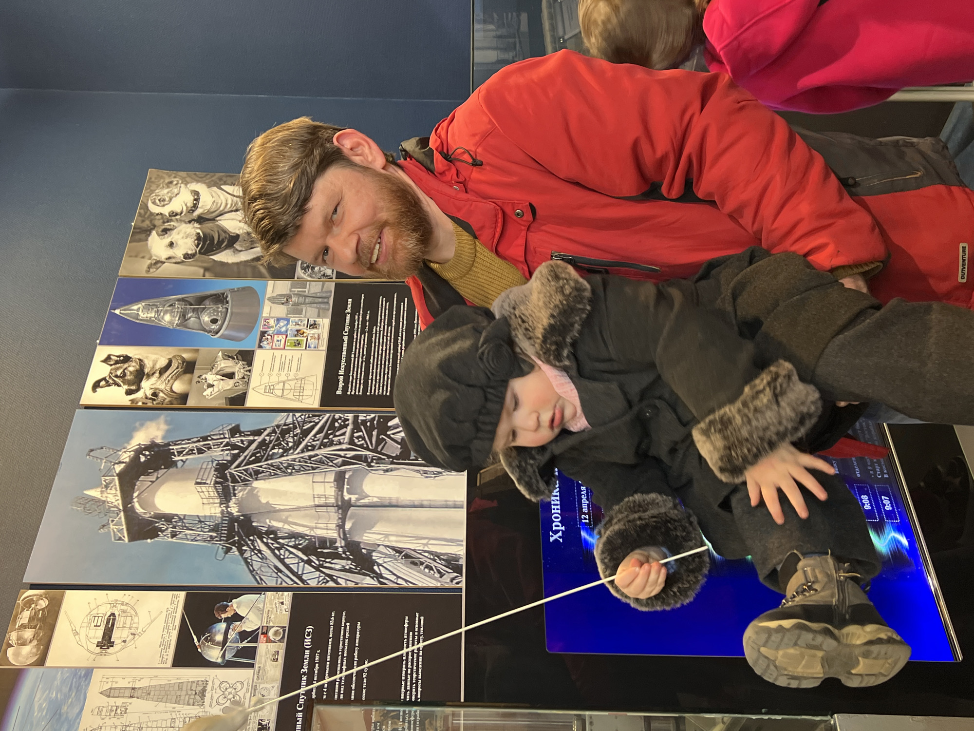 The image shows a man and a child, likely at a museum in Saint Petersburg, Russia. The man has a beard and is wearing a red jacket. The child is wearing a dark coat with fur trim and boots. They are positioned in front of several exhibits, including pictures and diagrams of rockets and spacecraft. The exhibits feature text in Russian, including phrases like "Искусственный Спутник Земли (ИСЗ)" (Artificial Satellite of Earth) and "Хроник" (Chronicle), as well as dates like "12 апреля" (April 12). The child appears to be using a pointer on a display screen that indicates the time of start for a rocket.