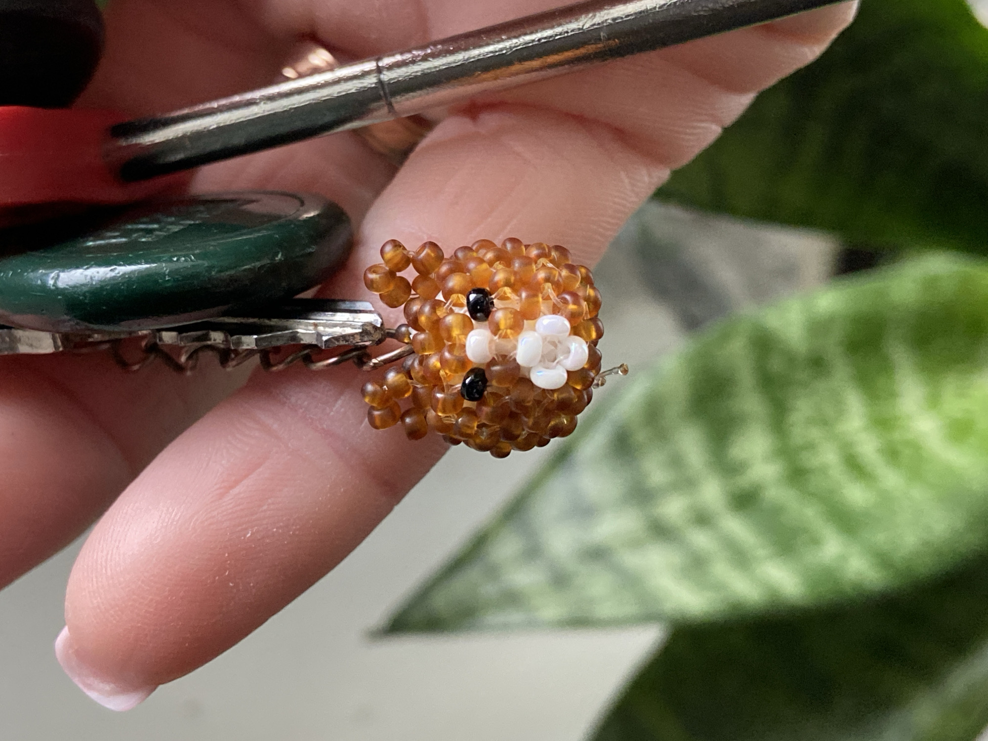 The image provides a close-up view of a person's hand holding a set of keys adorned with a beaded charm. The hand, with a visible light pink manicured fingernail on the ring finger, securely grasps the items. Several metal keys are present on a key ring, including one key distinguished by a dark green, possibly plastic or rubber, head.
The focal point is a small, intricately designed beaded charm. It is crafted from numerous tiny, round beads, predominantly in shades of amber and light brown, forming the body of what resembles a stylized animal, possibly a bear or hamster. The charm features two small black beads for eyes and a small cluster of white beads forming a snout or muzzle, with another black bead indicating a nose.
In the softly blurred background, the distinctive variegated green leaves of a plant, characteristic of a snake plant (Sansevieria), are visible, suggesting an indoor setting. The lighting appears to be natural daylight. No text is visible anywhere in the image.