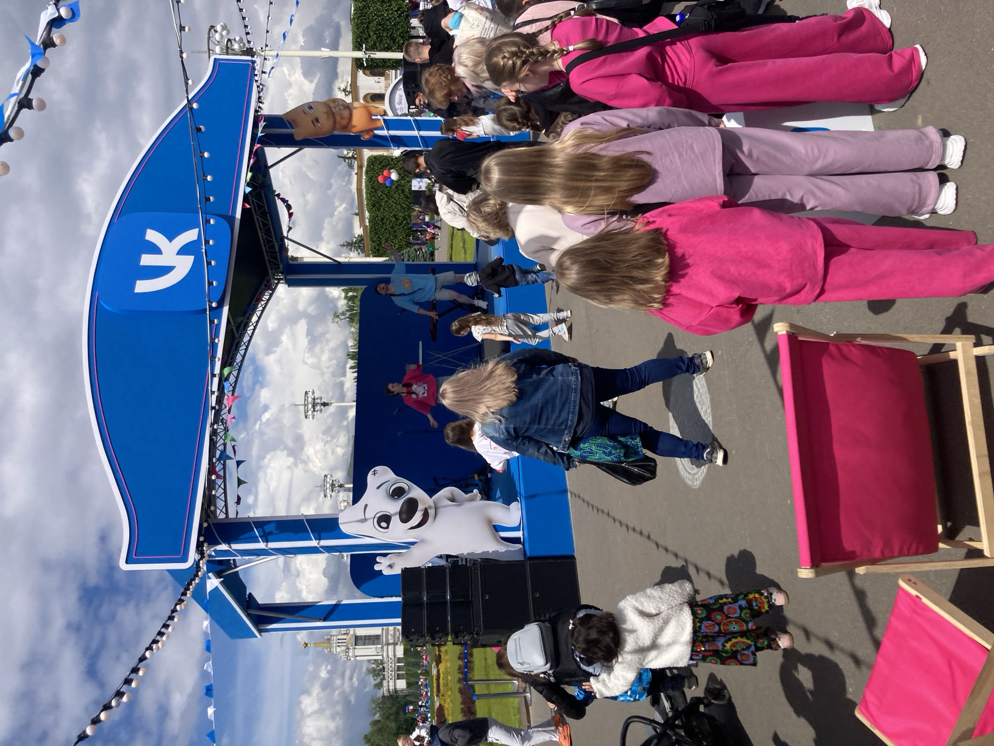 Captured outdoors during daylight hours, the image portrays a gathering of individuals in what is identified as Moscow, Russia. An open stage is the focal point, indicated by the presence of stage equipment and a blue backdrop emblazoned with a white "Y" logo, along with a mascot figure. Several individuals are performing on the stage. The audience is seated, with some individuals lying on mats or blankets and others standing, observing the activities. A variety of structures can be seen in the background, appearing to be part of a larger public space or park. The scene is lit by natural sunlight.