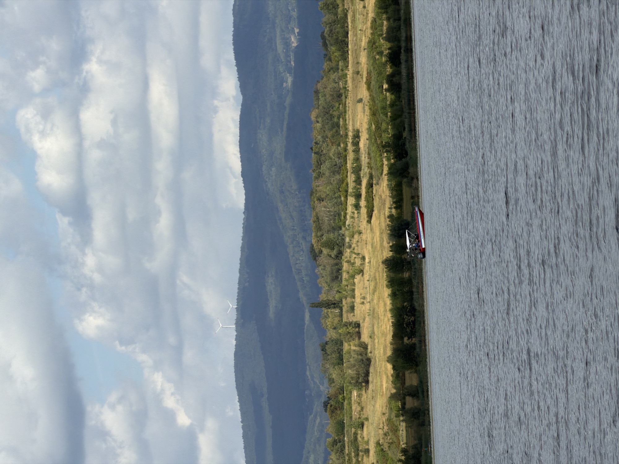 The image captures a scenic view of a lake with a small red and white boat on the water. In the background, rolling hills covered in lush green trees rise, with two wind turbines visible on the highest peak. The sky above is filled with dramatic, grey clouds interspersed with patches of blue. The terrain surrounding the lake appears to be a mix of grassy fields and dense foliage. While no specific landmarks confirm the location in Most, Czechia, the presence of wind turbines on hills is consistent with some landscapes found in the region. The overall scene suggests a tranquil, natural environment.