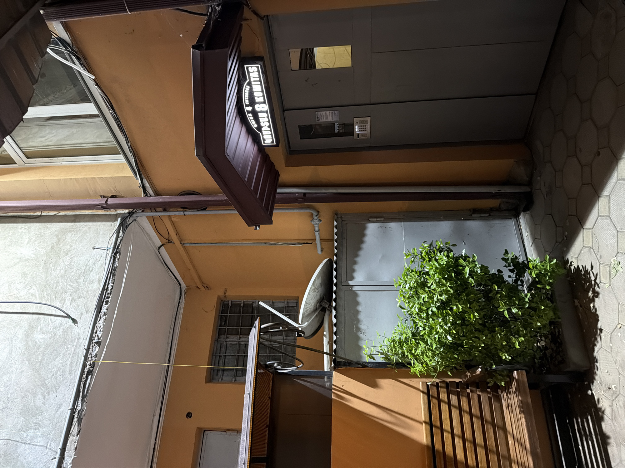 A low-angle view captures an outdoor alleyway in Yerevan, Armenia, at night. The structure is made of multiple buildings facing the camera with a complex layout and mixed uses. A dark awning with illuminated vertical text, "SARGSYAN KOMITAS," hangs above the street level. A large metal door dominates the upper level, with a smaller doorway beneath. A satellite dish is mounted, positioned in front of a barred window and a leafy green bush. The environment includes walls of grey stone and orange stucco, utility lines, and a combination of natural and artificial lighting.