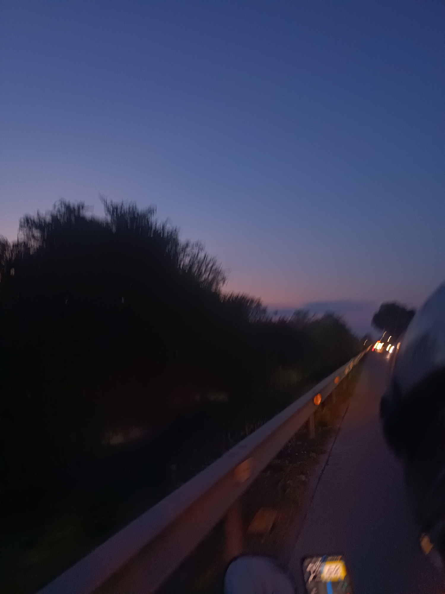 The image depicts a dusky scene from what appears to be a road in Hua Hin City Municipality, Thailand. A metal guardrail runs along the right side of the frame, illuminated by the low light. Beyond the guardrail, a dark, overgrown area with trees is visible. In the distance, there are faint hints of other vehicles' headlights. The sky transitions from a dark blue above to a faint orange glow on the horizon. A partial view of a person, possibly a motorcyclist, and a navigation device are in the foreground.