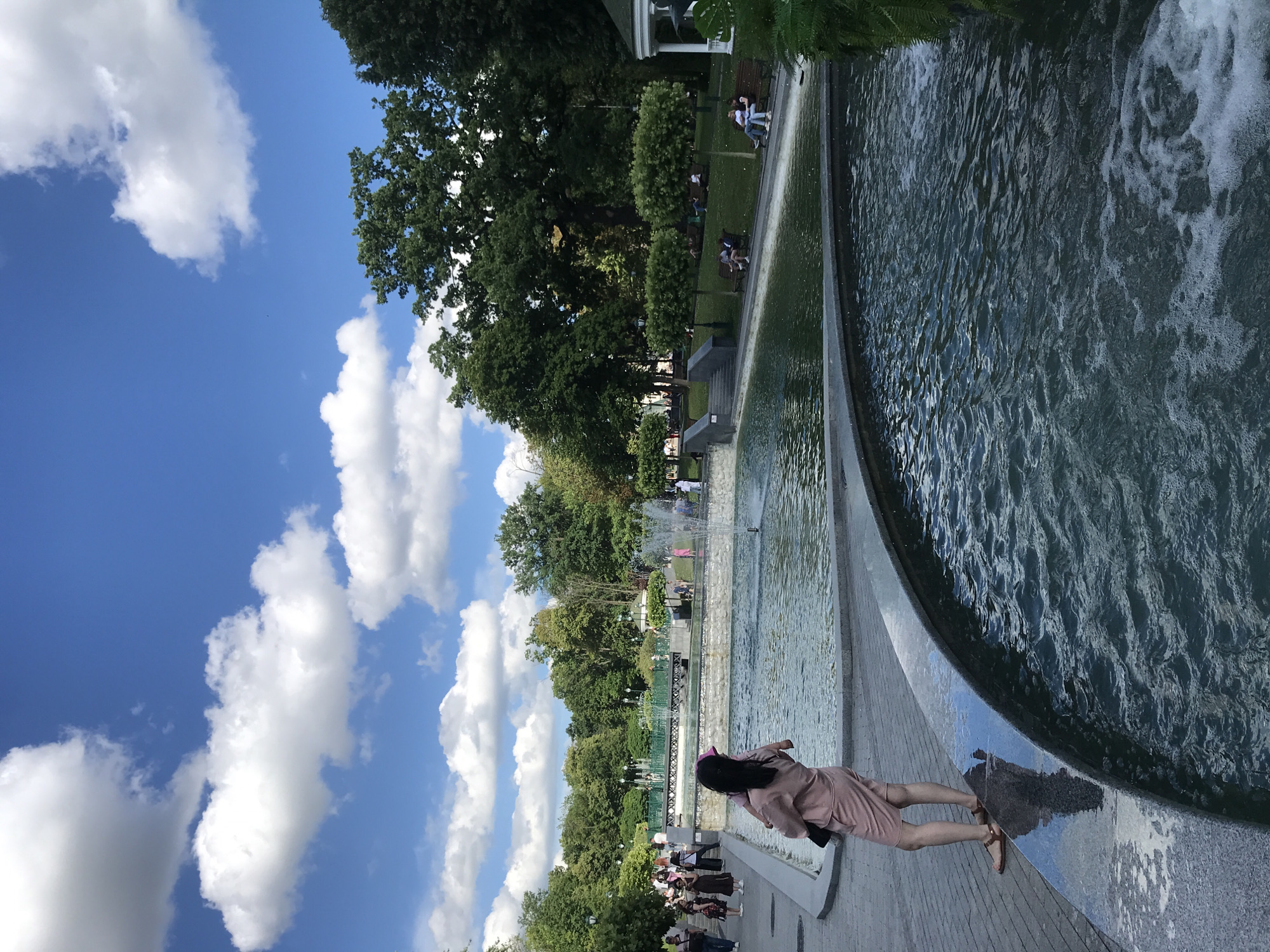This is an outdoor daytime image of a park in Kharkiv, Ukraine, featuring a multi-level fountain. In the foreground, a woman with dark hair is carrying a child on her hip, facing away from the camera. She is wearing a pale pink dress and sandals. The fountain's water is visible with ripples and some foam. The park itself has paved walkways, grass, benches, and mature trees. In the background, several people are seen walking and sitting on benches, and there are multiple jets of water spraying from the fountain. The sky is blue with scattered white clouds.