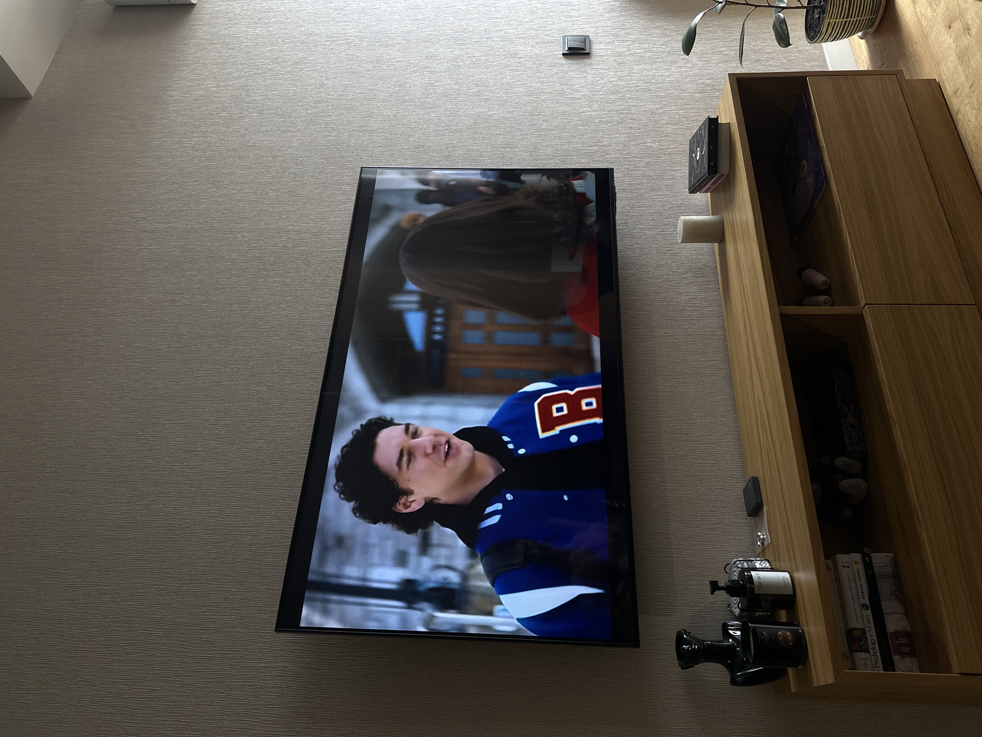The image shows a television mounted on a light beige wall. On the TV screen, a scene from a show is displayed, featuring a young man in a blue jacket with the letter "B" on it. Adjacent to the TV is a wooden cabinet with open shelves. On the shelves are various items, including books, decorative stones, and toiletries. The camera angle suggests a low perspective, looking up at the TV and cabinet. The environment suggests an indoor setting within a modern living space. The location in the city of Yerevan, Armenia is not discernible from the visual data.