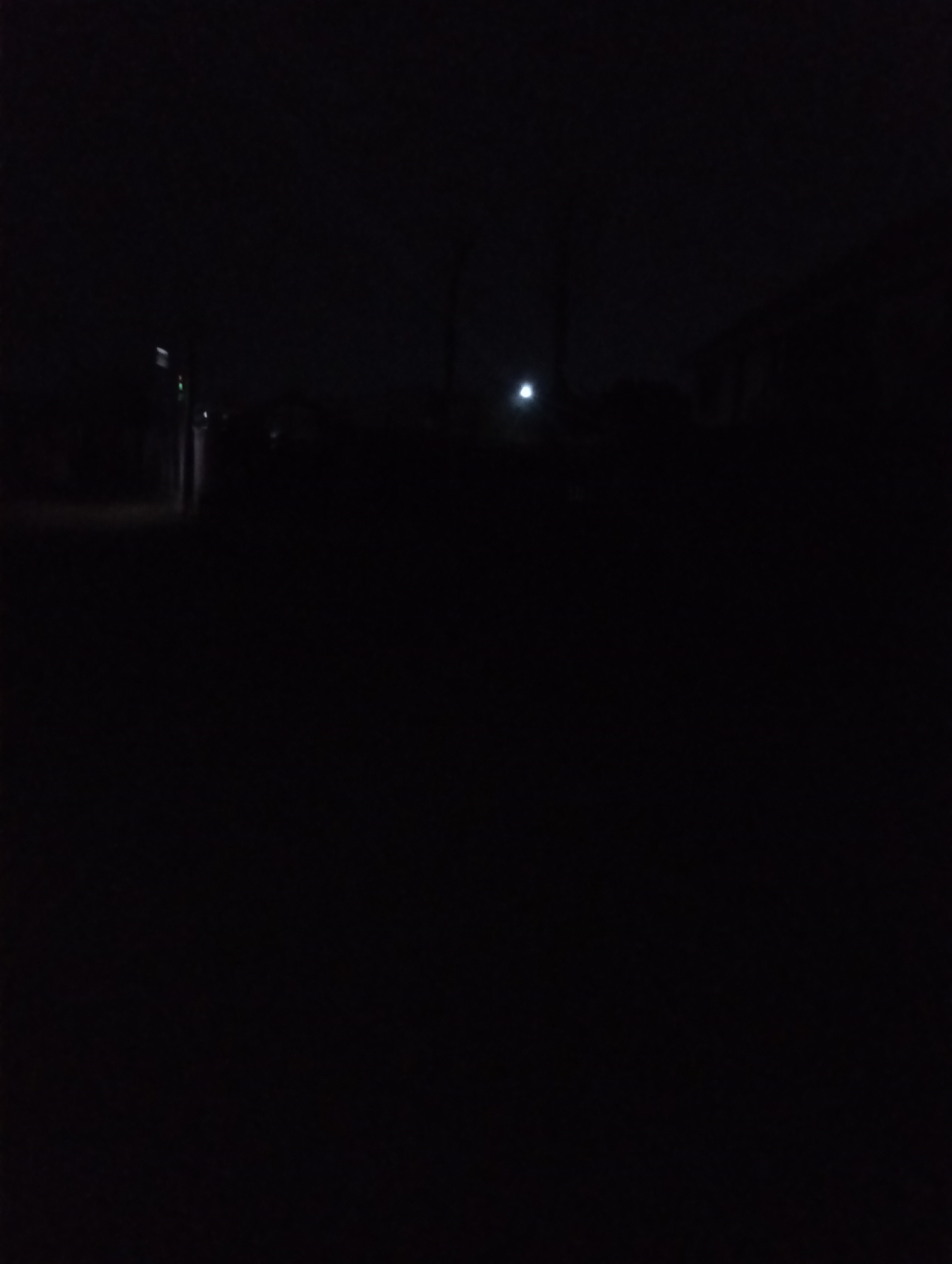 The image shows a very dark scene, likely at night. It appears to be a street in Jos, Nigeria, given the prompt's context. There are hints of buildings or structures on both sides of the street. A faint light source, possibly a street lamp, can be seen further down the road. The foreground is dominated by darkness, making it difficult to discern any details. There are no visible people or specific objects that can be identified with certainty due to the low light conditions.