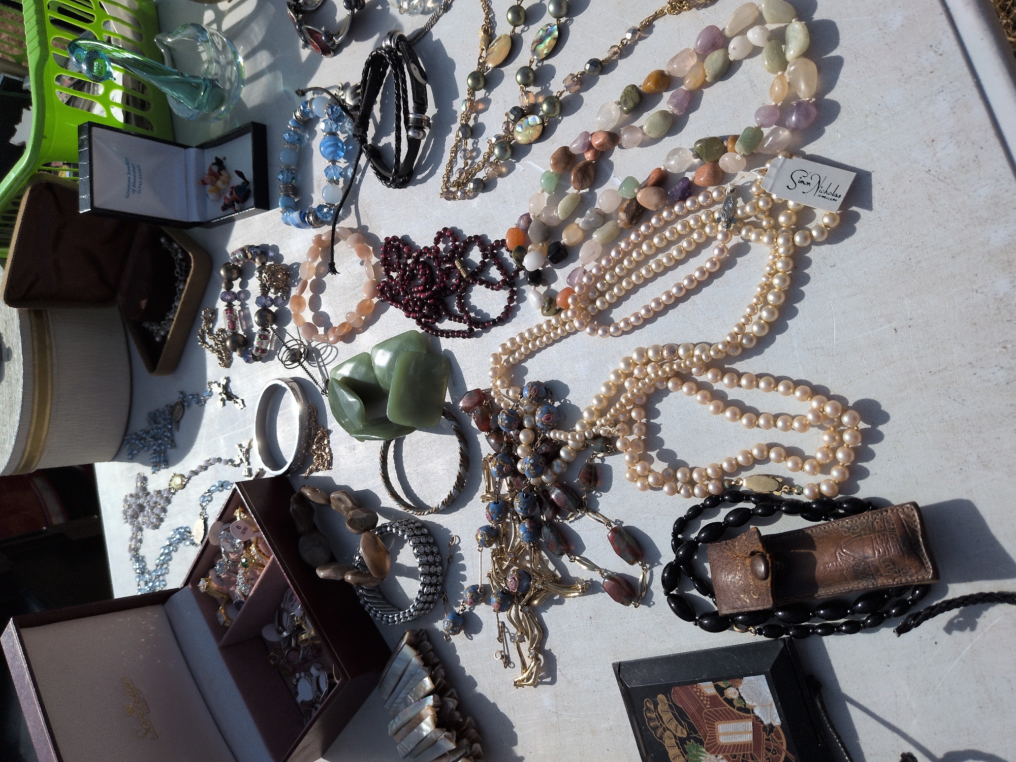 The image displays a collection of jewelry laid out on a white, possibly outdoor, surface. The items are predominantly necklaces, bracelets, and some earrings, suggesting a market stall or a personal sale.
Visible items include:
* A burgundy jewelry box, open, revealing various rings and other small trinkets.
* Several strands of pearls in different sizes and shades of cream.
* A beaded necklace with light blue and silver-colored beads.
* A necklace with dark red, garnet-like beads.
* Bracelets made of various gemstones, including jade-like green stones, amethyst, and other pastel-colored stones.
* A black leather pouch with a button closure, possibly for storing jewelry.
* A thin black bracelet with metallic accents.
* A silver-colored bangle.
* A set of mother-of-pearl shells, possibly part of a decorative item or jewelry.
* A small black box with a decorative oriental-style lid.
* A small glass sculpture of a bird.
* A green plastic basket in the background.
The lighting suggests it is daytime, with strong shadows indicating sunlight. There are no people visible in the frame. The surface appears to be a rough, textured material, consistent with a market table or a worn outdoor surface.
The image is undated and unlocated, but the presence of a diverse array of jewelry, along with a plastic market basket, strongly suggests a casual retail environment, such as a car boot sale or a flea market. The items themselves appear to be of mixed quality and style, ranging from what seems to be costume jewelry to potentially more valuable pieces.
Without further context or visual cues, pinpointing the exact location within Claverley, United Kingdom, is not possible. However, the overall scene is consistent with a typical outdoor market setting found in many rural or semi-rural locations within the UK.