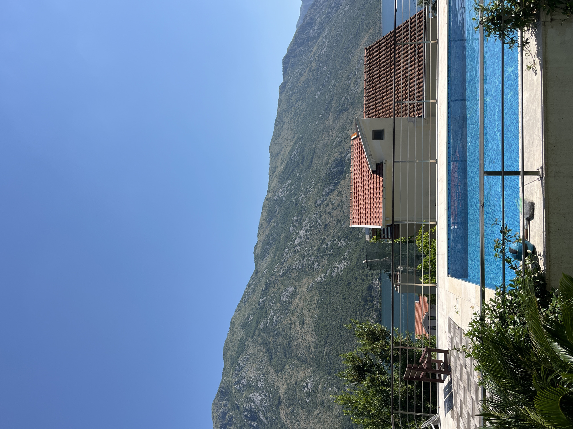 A clear, cloudless blue sky dominates the upper portion of the image, with a large, verdant, and rocky mountain range filling the middle ground. Below the mountain, a bright blue swimming pool with a tiled bottom reflects the sunlight, bordered by a stainless steel railing. To the left of the pool, a wooden bench sits on a tiled patio. Behind the pool, several houses with red-tiled roofs are visible. In the foreground, lush green foliage partially obscures the view. The scene appears to be a serene outdoor setting, likely a private residence or resort, overlooking a body of water and mountains, consistent with the picturesque scenery found in Perast, Montenegro.