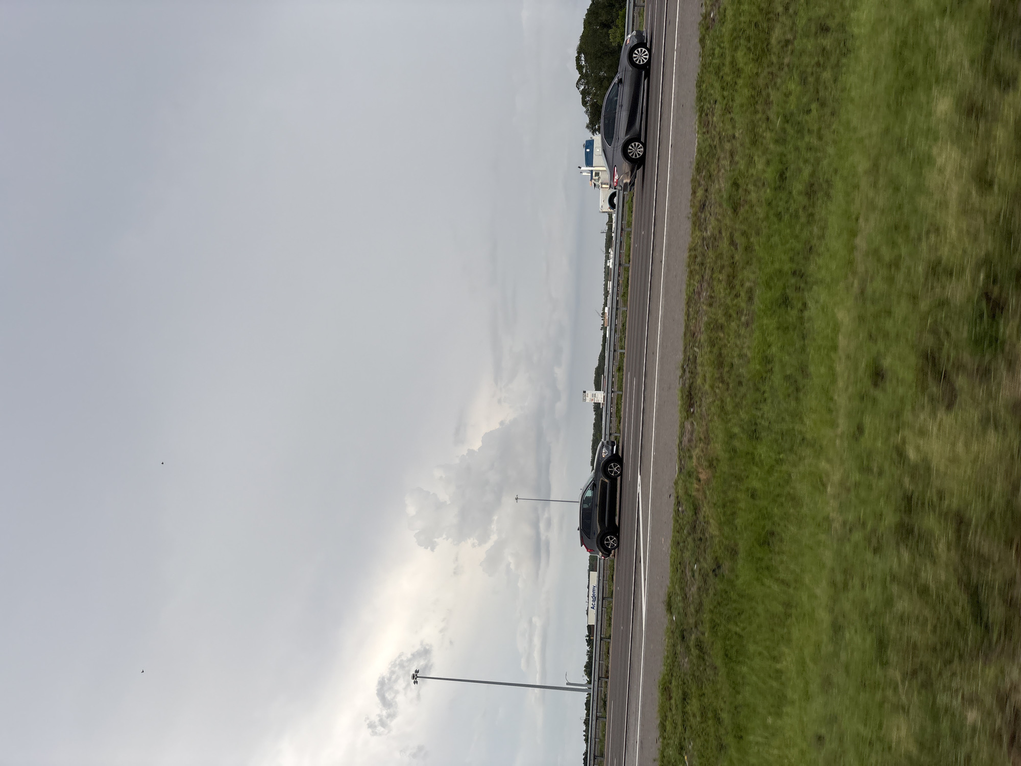 In an exterior, medium shot captured from a moving vehicle on an overcast day, two lanes of a highway are visible with a grassy median and shoulder. On the left side of the highway, a dark-colored SUV is traveling in the right lane. In the distance, a large white trailer truck is parked on the shoulder with a blue trailer. On the right side of the highway, a gray sedan is driving in the left lane. In the far distance, several trees and buildings are visible behind a guardrail. The sky is filled with gray clouds, and two small birds can be seen flying in the upper left quadrant of the image. The scene appears to be located in an urban or suburban area, possibly near an industrial zone, as indicated by the presence of the truck and trailer. The location is the city of Bradenton, United States.