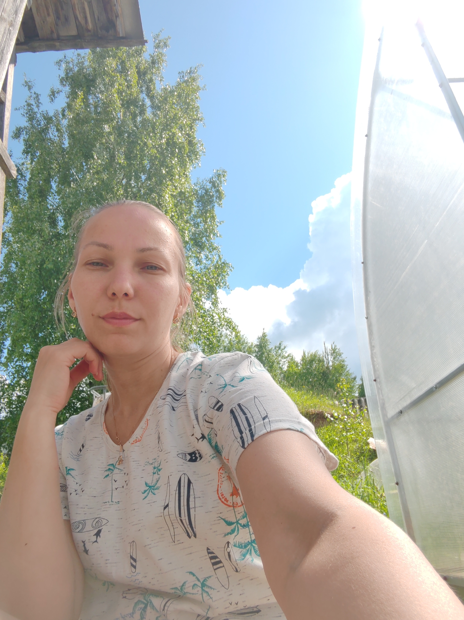 A woman is captured in a low-angle, outdoor selfie, with her head and upper torso filling the majority of the frame. She has fair skin, light brown hair pulled back, and is looking directly at the camera with a neutral expression. She is wearing a light-colored t-shirt with a repeating pattern of surfboards, palm trees, and other tropical motifs. Her left arm is bent, with her chin resting on her hand, and her right arm is extended forward, holding the camera.
The background is a bright, sunny sky with some fluffy white clouds. To the left, a tall green tree with dense foliage is visible, partially obscured by wooden beams from what appears to be a simple structure. To the right, a portion of a translucent, possibly polycarbonate, greenhouse structure is present. The overall impression is one of a casual, outdoor setting in a rural or suburban environment. The context suggests the photo was taken in Чухлэм, Russia, during daylight hours, under clear, sunny weather conditions.