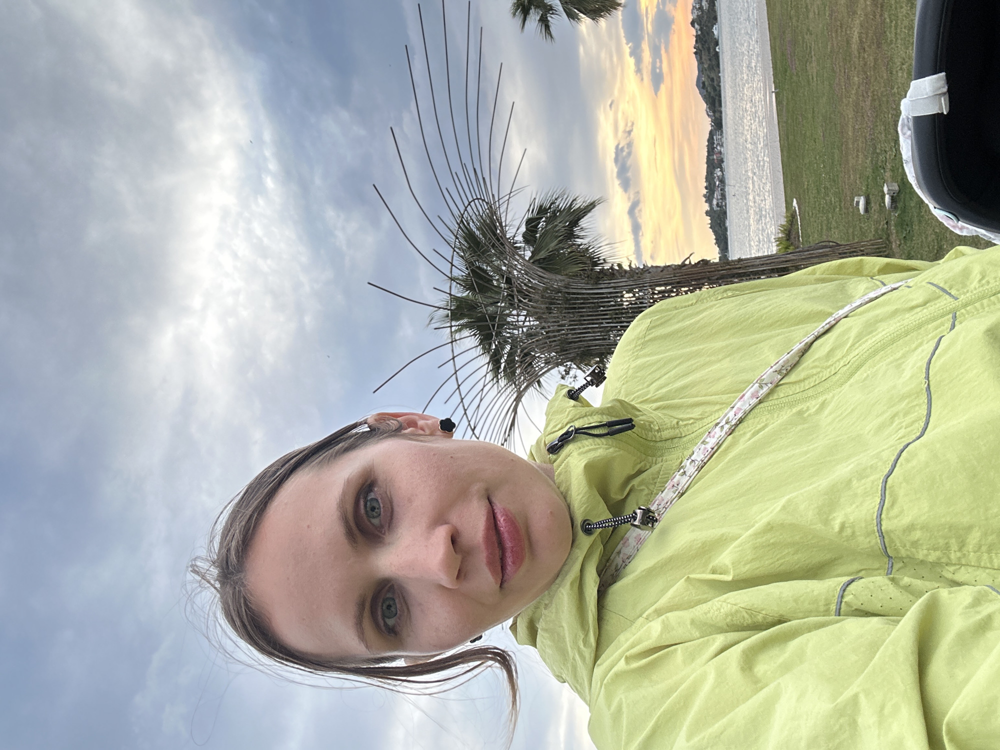 A selfie shows a woman in a chartreuse jacket. Behind her are a metallic sculptural tree with elongated metal branches, and a palm tree. In the background are a body of water, a coastline with buildings, and a sky with grey and yellow clouds. The location is likely in Fethiye, Turkey.