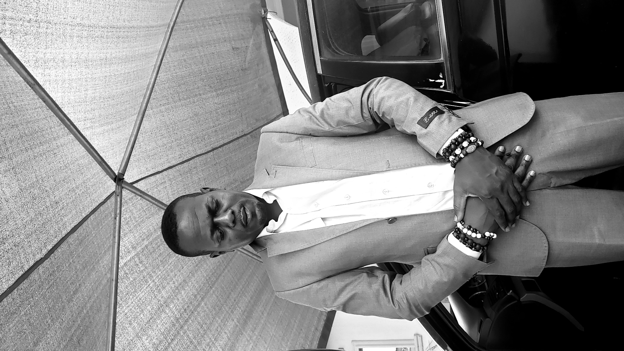 This black and white, portrait-oriented image features an adult man, likely in Sabon Tasha, Nigeria, captured in a posed shot. The man, with dark skin and short hair, stands or sits, facing slightly to the right with his head tilted back and eyes appearing to be closed or cast downwards, giving him a serene or contemplative expression.
He is dressed in a light-colored suit jacket over a white collared shirt, unbuttoned at the top. Both of his wrists are adorned with multiple beaded bracelets, predominantly black and white with some featuring patterned or metallic accents. His hands are clasped together in front of him. A small, distinctive label reading "Z ZARA" is visible on the left sleeve of his suit jacket.
To the right of the man, parts of a dark object, possibly a car door or its interior, are visible. Above and behind him, a large, light-colored canopy or tent-like structure provides shade, its textured surface and intersecting support poles prominently displayed. The overall setting suggests an outdoor or semi-outdoor location during the daytime.