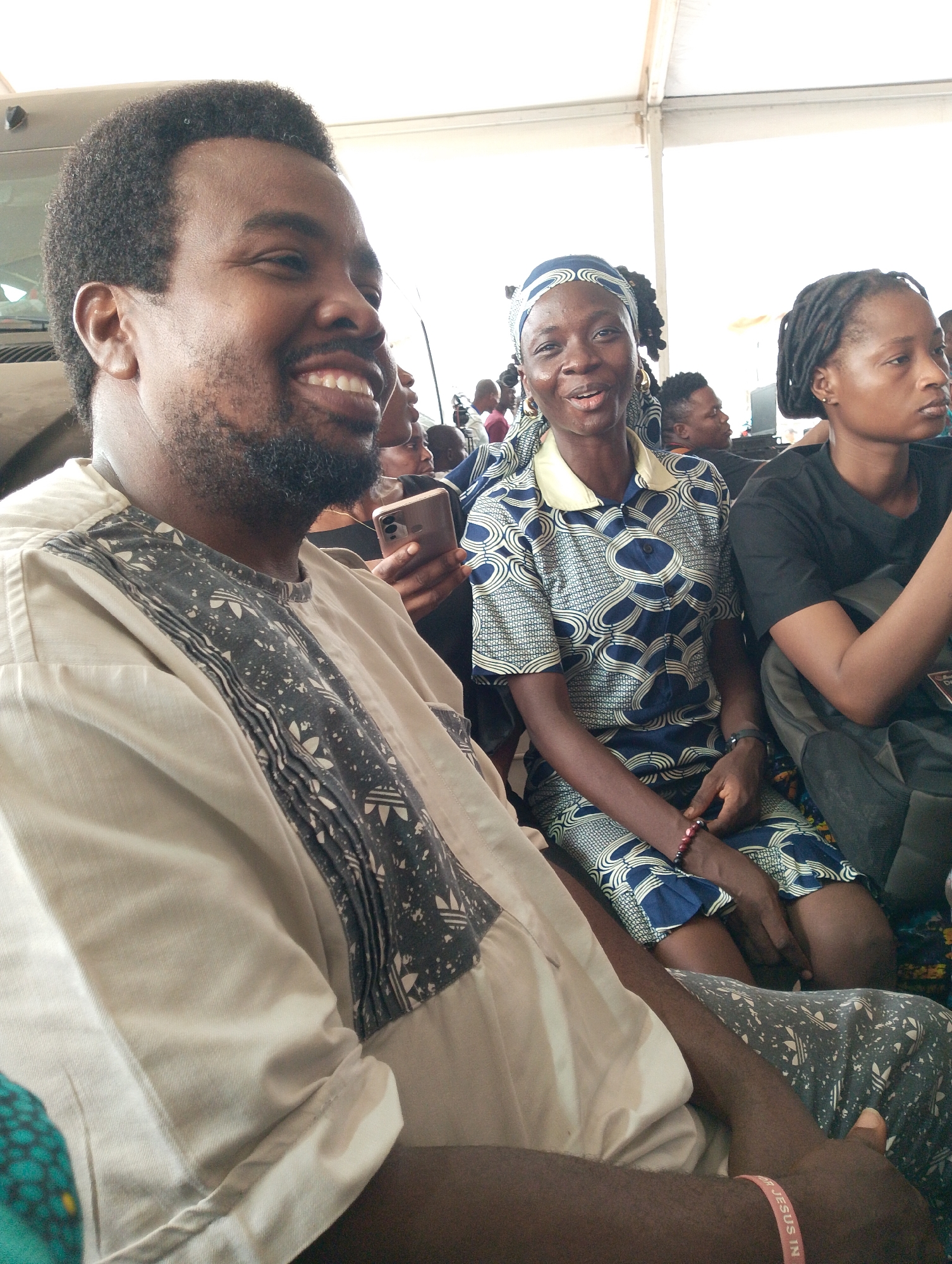 The image shows three people sitting closely together, with other individuals visible in the background, suggesting a crowded indoor location, possibly a tent. The photograph might be taken in the city of Dushepe, Nigeria, due to the traditional attire of the individuals and the presence of several people in the background.
A man in the foreground is smiling and wearing a tan and black patterned shirt, while a woman next to him is wearing a blue and white patterned outfit with a headscarf and also smiling. To her right, another woman in a black t-shirt is looking away from the camera with a more neutral expression. The lighting appears bright and even, suggesting a well-lit environment, likely due to natural light from the tent’s opening. One individual is using their cell phone.