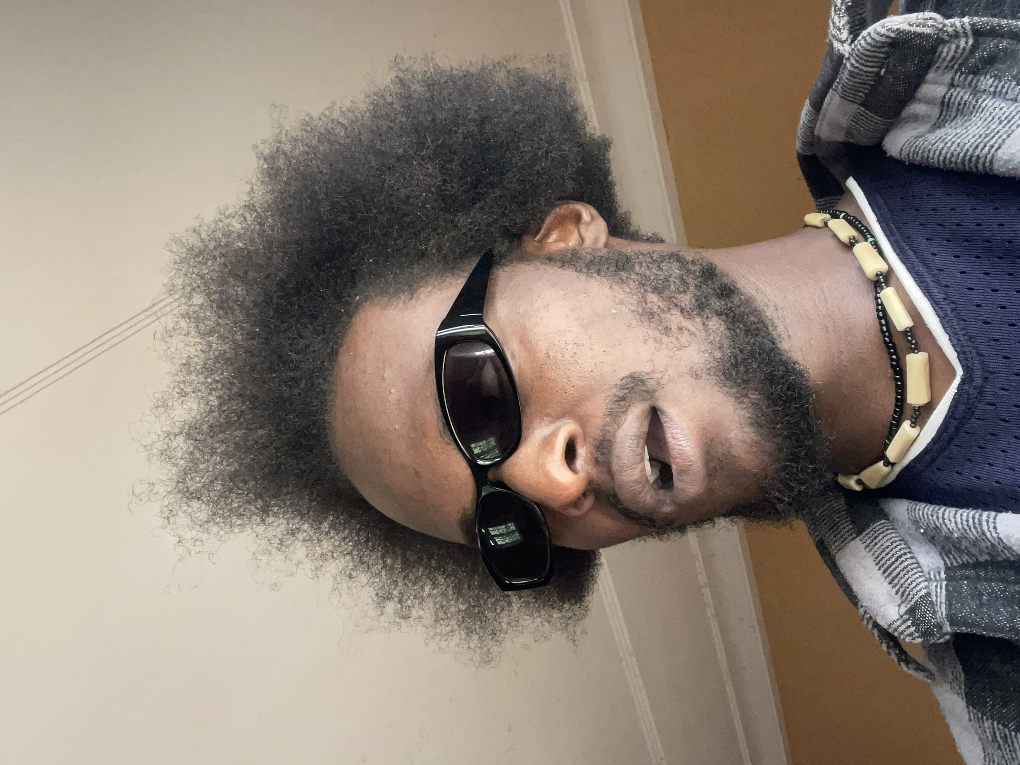 The image shows a close-up, head-and-shoulders portrait of a Black man wearing sunglasses, a plaid shirt, and a beaded necklace. The man has an afro hairstyle and a beard. He is looking slightly to his left, with his mouth open. The background appears to be a room with a light-colored ceiling and a yellow-painted wall. There are no visible contextual clues to indicate the specific location within the city of Jos, Nigeria.