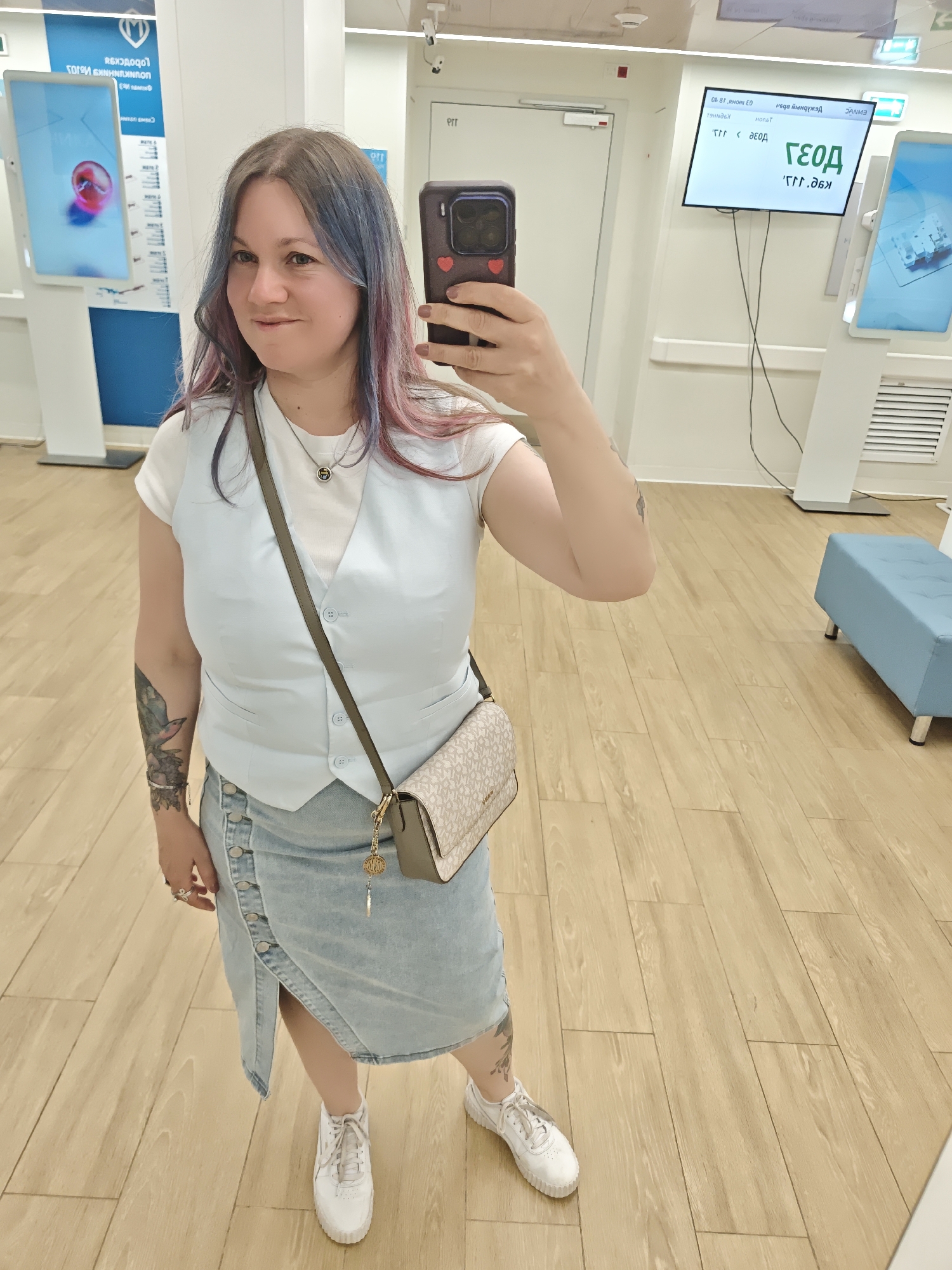 A woman takes a selfie inside what appears to be a modern, well-lit public service center or bank branch, possibly in Moscow, Russia. She is looking directly into the camera with a slight smile.
The woman has light brown hair with prominent blue and purple highlights. She is wearing a white short-sleeved t-shirt layered under a light blue sleeveless vest, a light blue denim midi skirt with a button-front and an asymmetrical hem, and white platform sneakers. She accessorizes with a silver-colored necklace featuring a circular pendant, several rings on her fingers, and a light-colored shoulder bag with a patterned exterior and a circular gold charm. A detailed tattoo of a bird and flowers is visible on her left forearm, and another smaller tattoo is on her right upper arm. She is holding a smartphone in a dark-colored case with a red heart sticker on the back.
The background shows a brightly lit interior with light wooden floorboards. To the left, there's a tall, white digital display stand with a screen showing a mirrored reflection of a logo (resembling Sberbank or MTS) and Russian text, including "СБЕРБАНК" (Sberbank) and "БАНКОМАТЫ" (ATMs). To the right, another large digital screen on the wall also displays mirrored Russian text. Key visible reversed text fragments on this screen include "04:81 НОЯБРЬ ЕО" (04:81 November EO), "ДЕЖУРНЫЙ ИНФОРМ" (Duty Information), "ОКНО" (Window), "ТАЛОН" (Ticket/Token), and "ГЕОД" with a code "ГГГ.ЭОД" below it, likely indicating a queue system or service point. A light blue upholstered bench with metallic legs is visible in the far right background. The overall setting suggests a customer service area or waiting room.