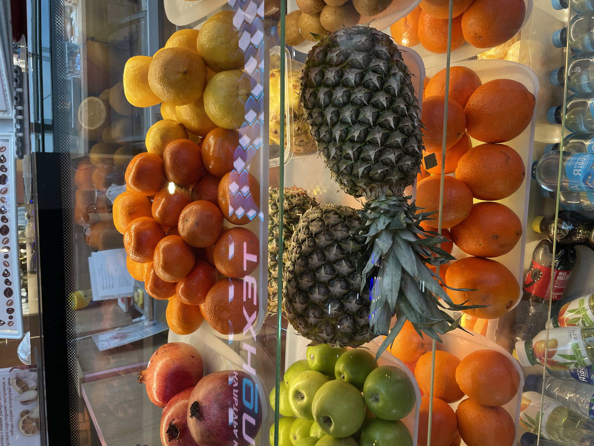 Here is a description of the image:
A slightly canted, eye-level indoor shot depicts a refrigerated display case filled with fresh fruits. The case is divided into sections by clear dividers. The fruits are arranged mostly in piles.
From left to right, there are rows of yellow citrus fruits and oranges, followed by two pineapples. Next to the pineapples are more oranges and green apples. The bottom left corner displays pomegranates. The right side of the display also contains bottled drinks, including water and juice.
The image also captures reflections on the glass of the display case. A poster with food items is visible on the left. The photo appears to have been taken in Kommunarka, Russia, as indicated by text on some signage inside the case. The image shows an array of fruits and beverages, typical of a store display.