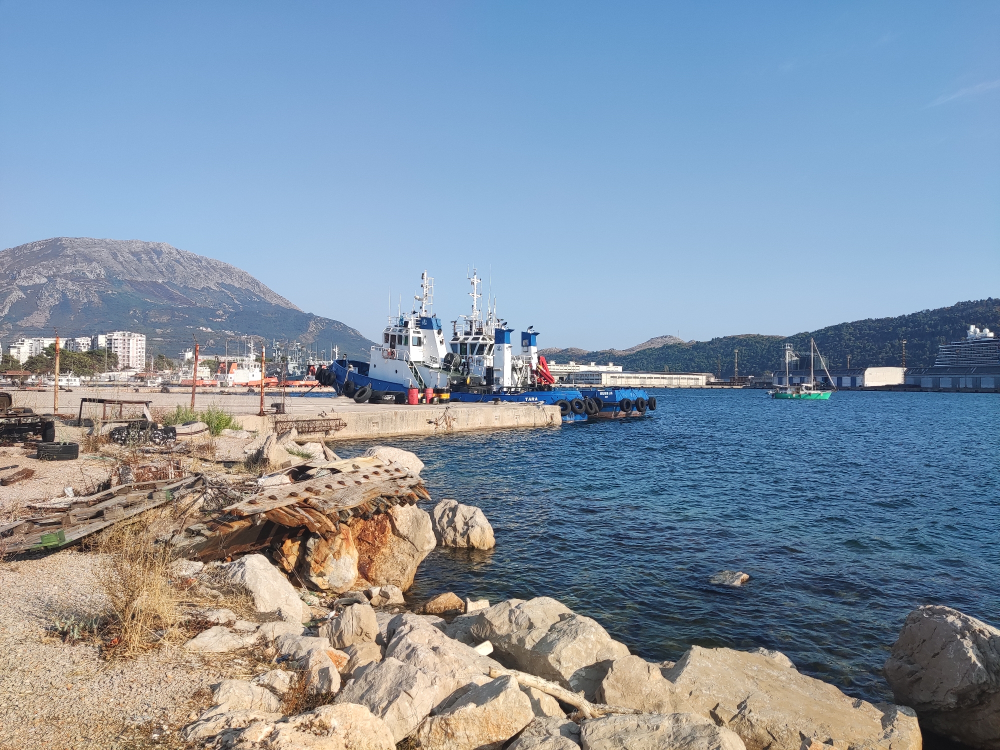 The image depicts a coastal scene in the city of Bar, Montenegro, likely in or near the port area. In the foreground, a rocky shoreline is visible, with a collection of weathered wooden debris and large stones. Beyond the shore, the blue waters of the Adriatic Sea are calm, with a few scattered rocks visible beneath the surface.
Dominating the middle ground are several tugboats docked at a concrete pier. These boats are white with blue accents and have various structures on their decks, including cabins and antennae. One of the tugboats is labeled "TARA." In the background, a range of buildings, including apartment complexes, line the coast. Further in the distance, a large, rugged mountain range rises behind the city.
The sky above is clear and a bright blue, indicating fair weather. The overall impression is of a working port with a backdrop of natural landscape, under a clear, sunny sky. There are no people visible in the image. The scene is static and peaceful, with the focus on the maritime activity and the surrounding environment.