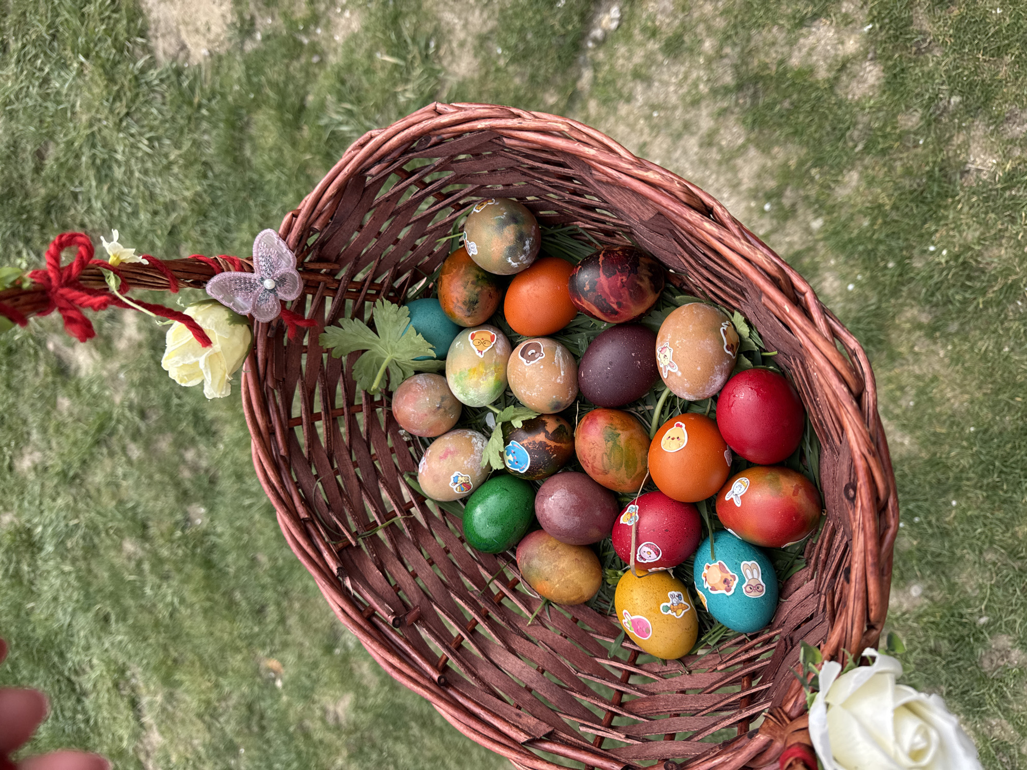 A high-angle shot shows a brown wicker basket filled with an assortment of Easter eggs. The basket is adorned with a red ribbon, a butterfly embellishment, and white roses. The eggs inside are painted in various colors, including brown, orange, red, green, and blue, some decorated with small stickers of animals and characters. The basket rests on a background of green grass. The location is in Sofia, Bulgaria, based on the origin of the image.