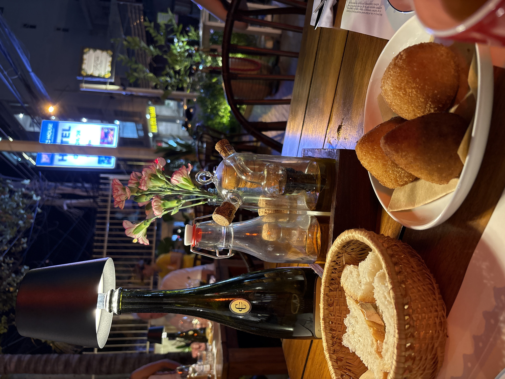 The image depicts an outdoor dining scene at night, likely a restaurant or cafe, situated in Nha Trang, Vietnam, with an Ibis Hotel sign visible in the background. The foreground features a wooden table presenting a meal.
On the table, a woven basket contains several slices of white bread. Adjacent to it, a white oval plate holds three golden-brown, breaded, fried food items, one conical and two spherical, each resting on a small piece of brown parchment paper. A portion of a red cup is visible on the right.
In the midground, a dark green bottle, possibly a wine bottle with a gold "E" or "C" emblem, is repurposed as a table lamp base, topped with a black lampshade emitting a downward light. Next to it, a small bouquet of pink and white flowers (likely carnations) is arranged in a clear glass vase. A wooden caddy holds three glass cruets: one with a red-and-white flip-top stopper containing a light amber liquid, and two others, one with a cork stopper, holding different amber and dark liquids, possibly oil and vinegar. Beyond these items, a neatly folded white napkin containing silverware is visible on a paper placemat.
The background is dimly lit and out of focus, showing other patrons seated at additional wooden tables. Visible street elements include lush green foliage, a metal fence, and blurry lights from passing vehicles. Overhead, multiple illuminated signs are present, including a prominent blue and white vertical sign displaying "IBIS HOTEL" and "HOUSE," indicating an Ibis Hotel nearby. Another smaller, square sign with yellow text is also visible. The overall scene suggests a casual evening dining experience.