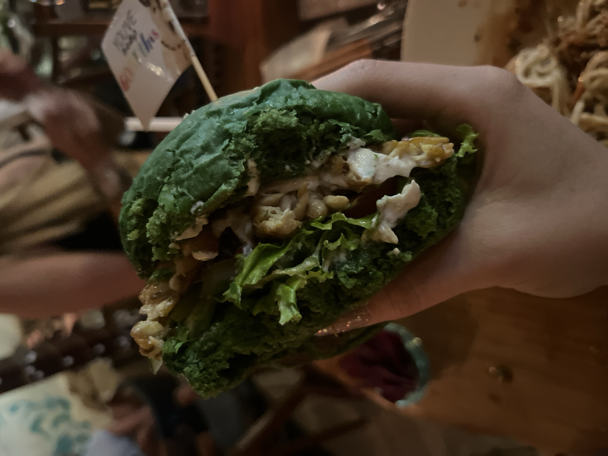 A close-up shot depicts a person holding a green burger in their hand. The burger appears to be filled with lettuce and a white sauce-like substance, along with fried pieces. A small white flag with writing on it is inserted into the top of the burger. The scene is indoors and also includes the background details such as food and people sitting at the table. Based on context, the location is likely in the Ubud District, Indonesia.