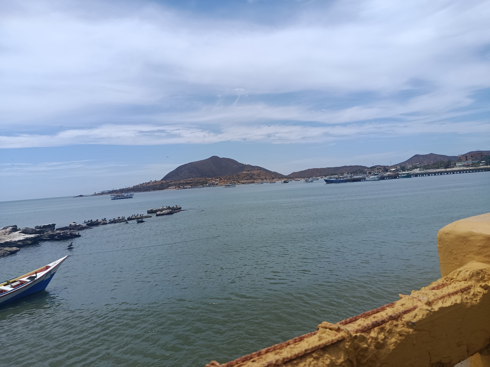 The photograph depicts a seascape in Juan Griego, Venezuela. The calm water extends from the foreground to the midground, where several boats are anchored, some near the shore. Rocky formations jut from the water, hosting groups of birds. In the distance, a mountainous landscape frames the horizon under a partly cloudy sky. A small fishing boat is visible in the bottom left corner of the image, and a weathered yellow concrete barrier occupies the bottom right corner.