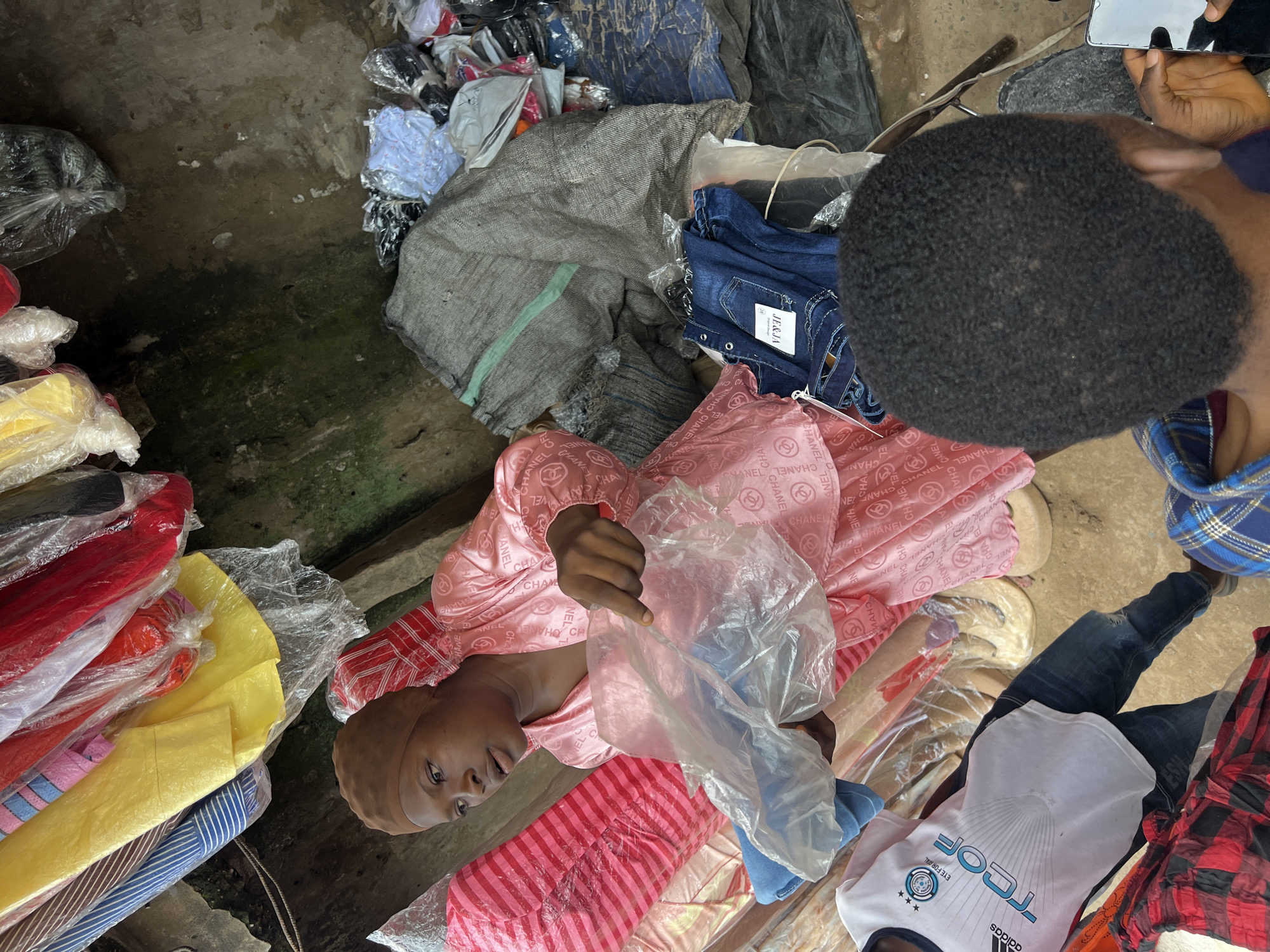 At a street market in Nkpor, Nigeria, a young Black woman wearing a brown head covering and a pink satin robe is seen behind a stall filled with various clothing items. She holds a transparent plastic bag containing blue denim jeans. Several racks of colorful clothing, some still in plastic wrapping, hang above her. To her right, there are piles of clothing, including more denim jeans, a large grey sack, and various other garments. In the foreground, the back of another person's head, also Black with short, curly hair, is visible, and they are holding a smartphone. The ground appears to be dirt or concrete, and the surrounding environment suggests a bustling outdoor market setting.