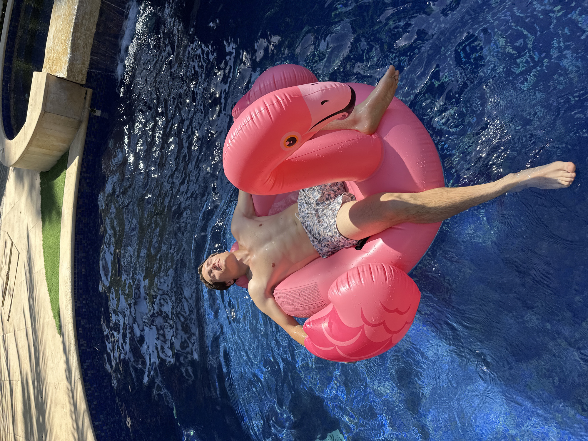 In the city of Savyon, Israel, a young man with light brown hair is relaxing on a pink inflatable flamingo float in a deep blue swimming pool. He is lying on his back with his eyes closed, enjoying the sunlight. The pool has a tiled edge, and surrounding the pool is a light-colored stone patio with palm tree shadows cast upon it. To the upper right, there is a curved stone structure with a small waterfall cascading into the pool, suggesting a hot tub or spa feature. The man is wearing patterned swim trunks. The water is clear and reflects the sunlight, creating a sparkling effect.