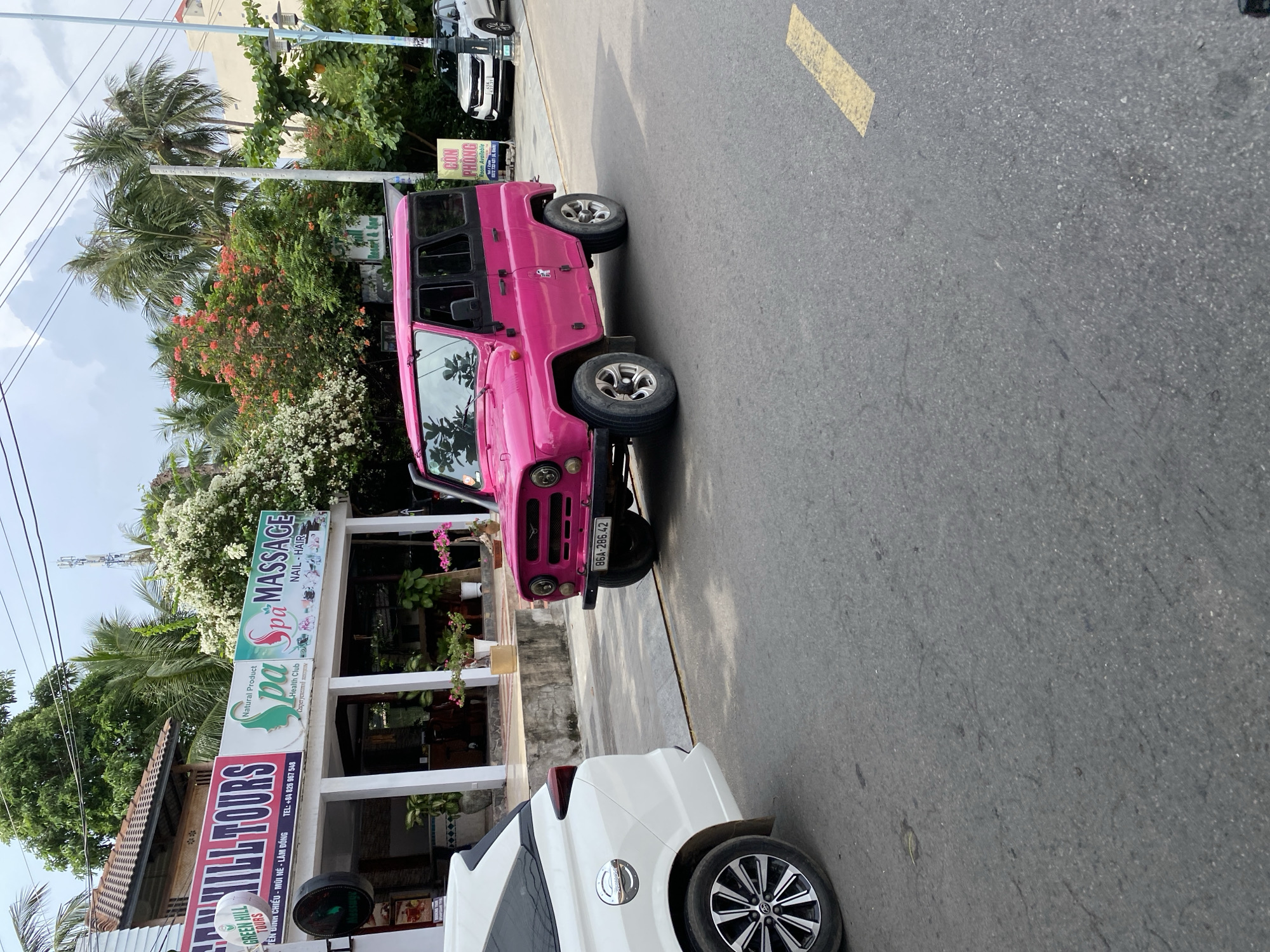 The image depicts a street scene in Phu Thuy Ward, Vietnam. A vibrant pink, boxy SUV, possibly a UAZ-469, is parked on the right side of the road. Its license plate reads "86A-286.42". In the foreground, the rear passenger side of a white SUV is visible on the left. Behind the pink vehicle, a white building with signs for "GREEN HILL TOURS" and "pa Spa MASSAGE" is situated. The building's facade is white with arched entrances and is lined with potted plants. Lush green trees and flowering bushes, some with pink and white blooms, surround the building. Further down the street, another white SUV is parked near a sign that says "CON PHONG Room Available". Power lines crisscross the sky, and the road is asphalt with a yellow painted line. The weather appears to be overcast.