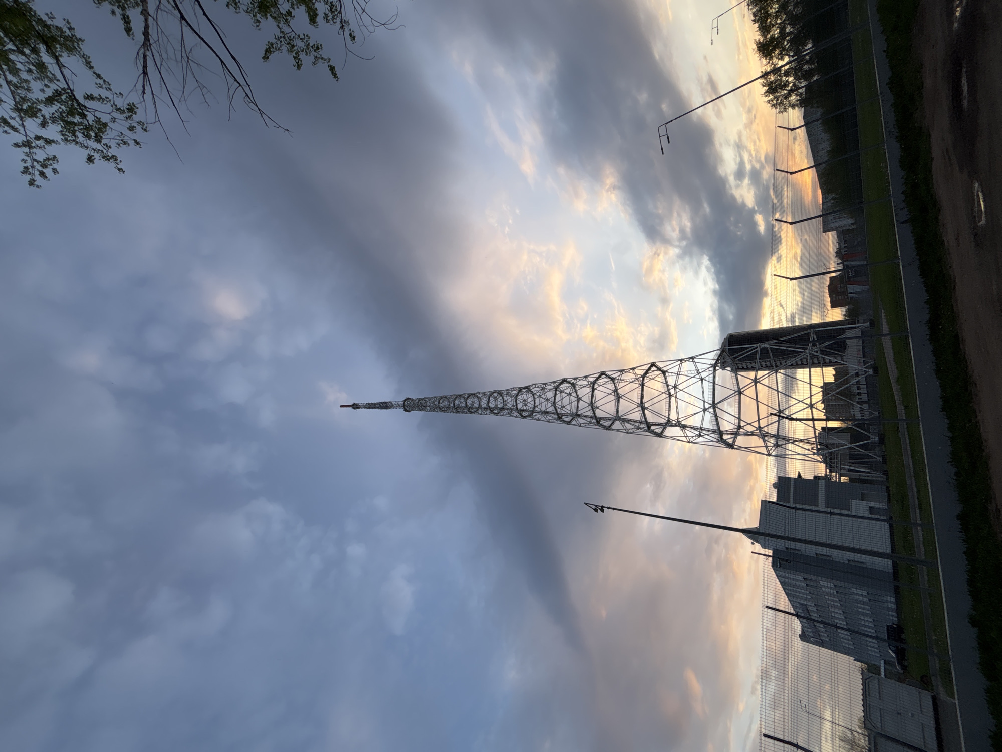 The image shows a vertical view of a radio tower in Perm, Russia, against a cloudy sky at sunset. The tower is a slender, lattice-like structure that tapers towards the top. The base of the tower is surrounded by buildings and a fence. A row of streetlights is positioned alongside the road. The sky is a mix of blue, gray, and orange from the setting sun. In the top-left corner, some tree branches and leaves are visible.