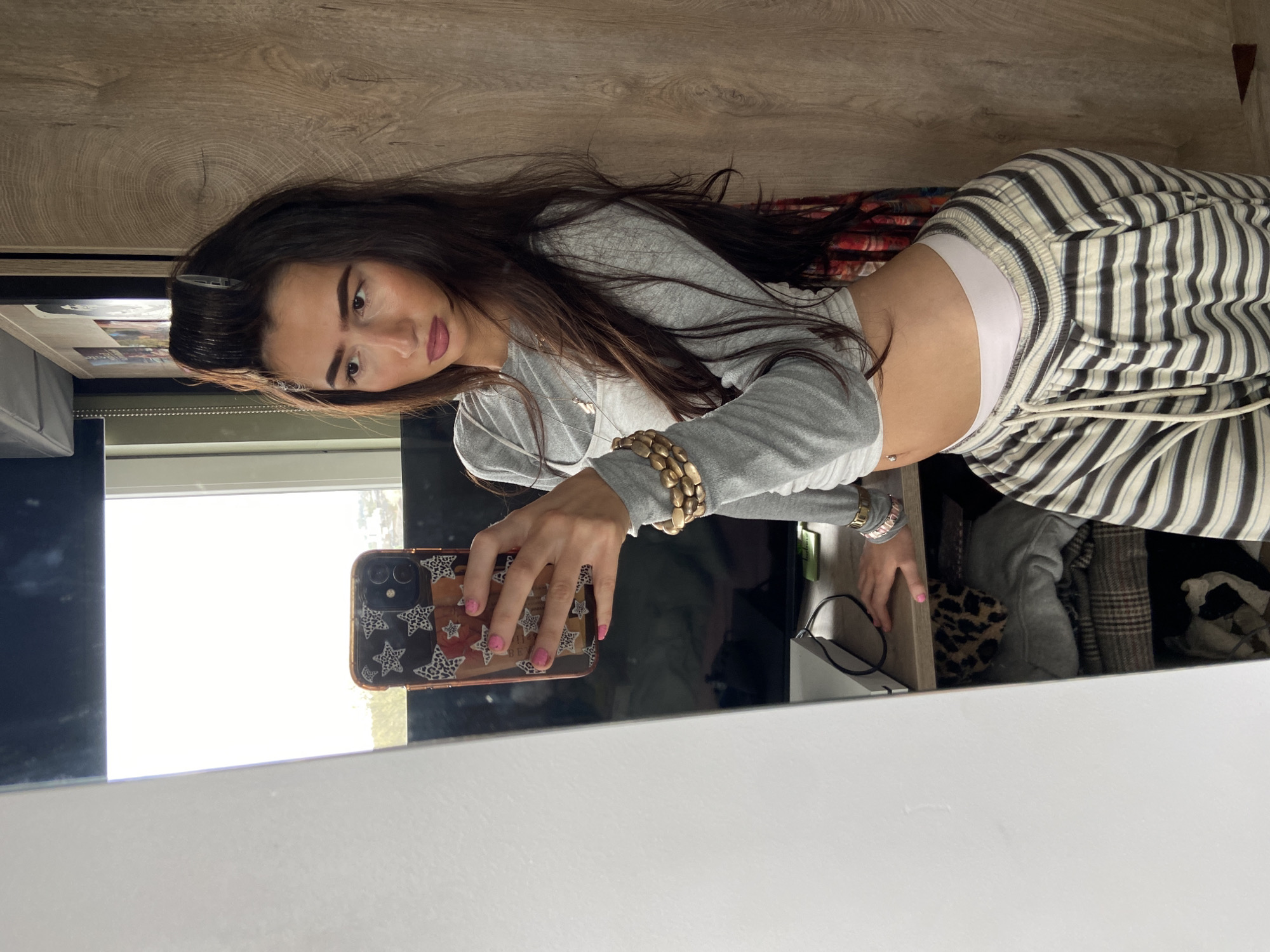 A young woman with long brown hair takes a selfie in a mirror. She wears a gray long-sleeved shirt, striped pants, and a white undergarment. Her phone has a patterned case and pink nail polish is visible. She has a hair clip in her hair and bracelets on her wrist. The reflection shows a room with a light-colored wooden surface, a window, and a cluttered area containing clothes and accessories. The location is not discernible as London, United Kingdom, without additional contextual clues.