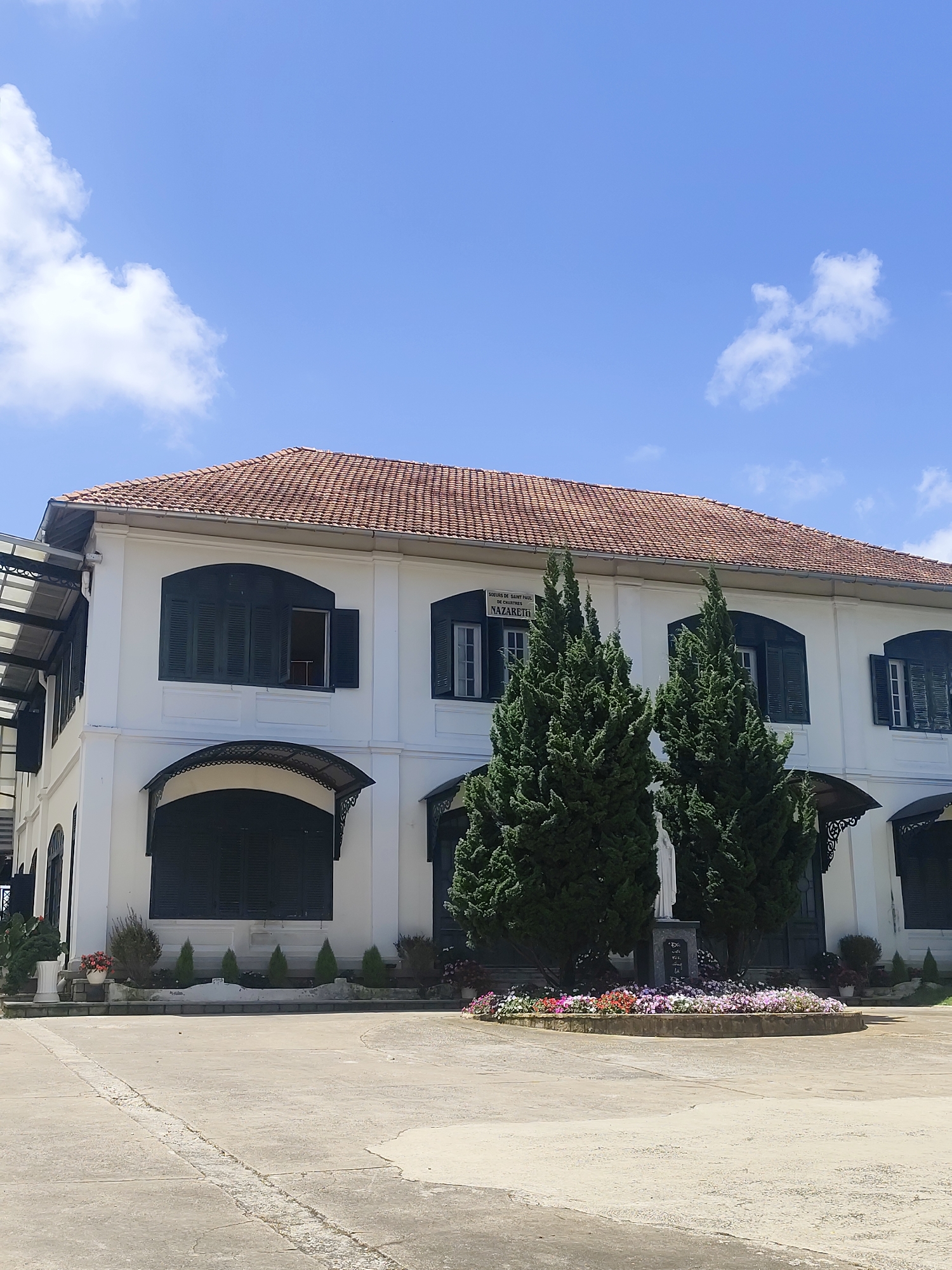 This image depicts a two-story white building with a red tile roof, situated in Da Lat, Vietnam. The building features dark green shutters on its windows, some of which are open, revealing glimpses of the interior. Above the entrance of the building, a sign reads "SOEURS DE SAINT PAUL DE NSTRUCTION DE NAZARETH". In front of the building, there is a paved courtyard and a circular flower bed containing a variety of colorful flowers. Behind the flower bed, two tall, slender evergreen trees stand, with a white statue of a figure, likely religious, positioned between them. Additional potted plants and small, conical evergreen trees are arranged along the facade of the building and the edge of the courtyard. The sky above is a clear blue with a few scattered white clouds. The overall scene suggests a serene and well-maintained environment, possibly a school or convent.