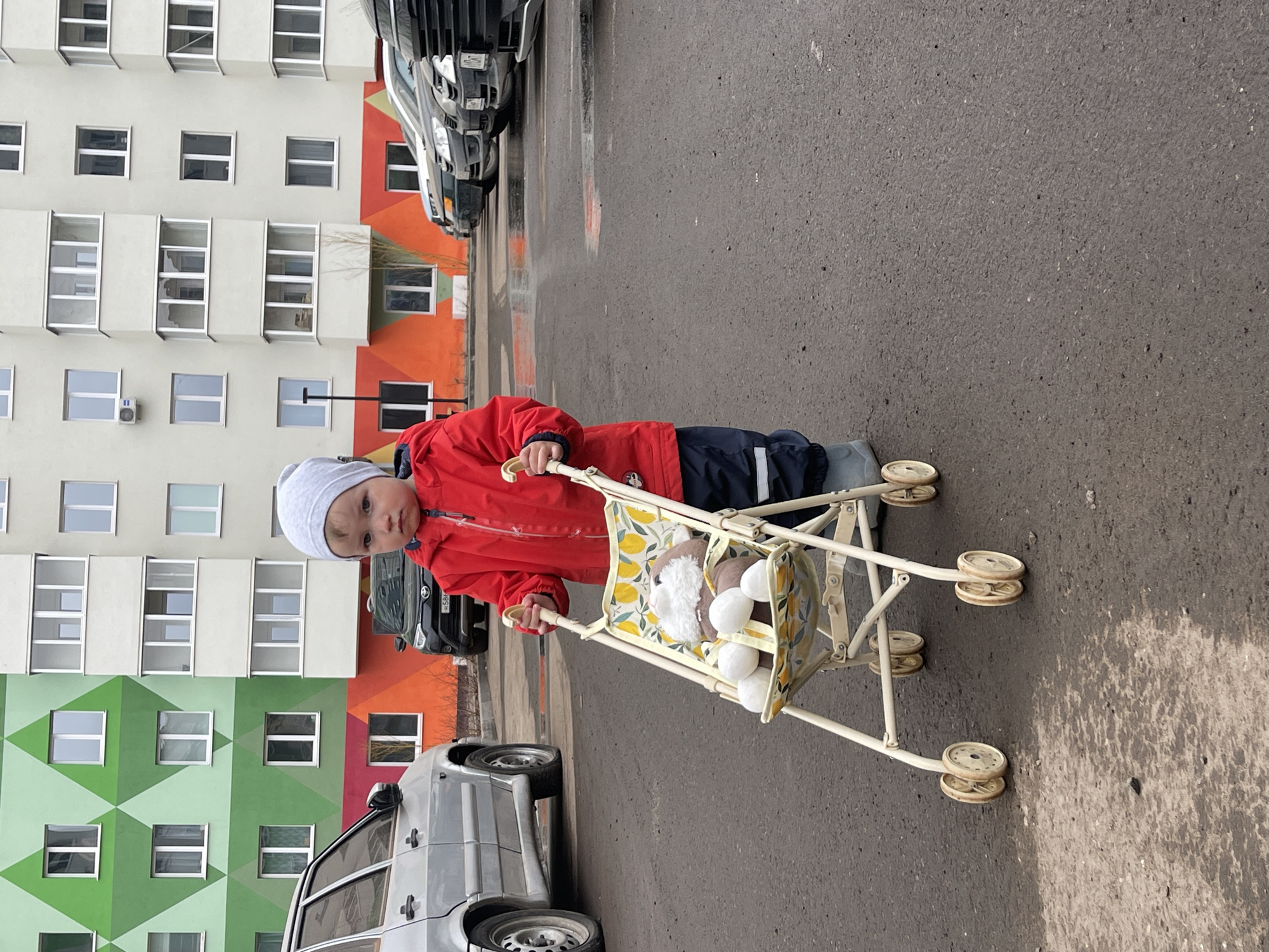 A child in a red jacket and gray hat stands on an asphalt surface, pushing a doll stroller. The stroller contains a stuffed animal, a brown and white fox with a white tail. Behind the child, a multi-story apartment building with windows is visible. The building's facade is decorated with green, orange, and white sections. Cars are parked along the side of the road next to the building. The scene is located in Izhevsk, Russia.
