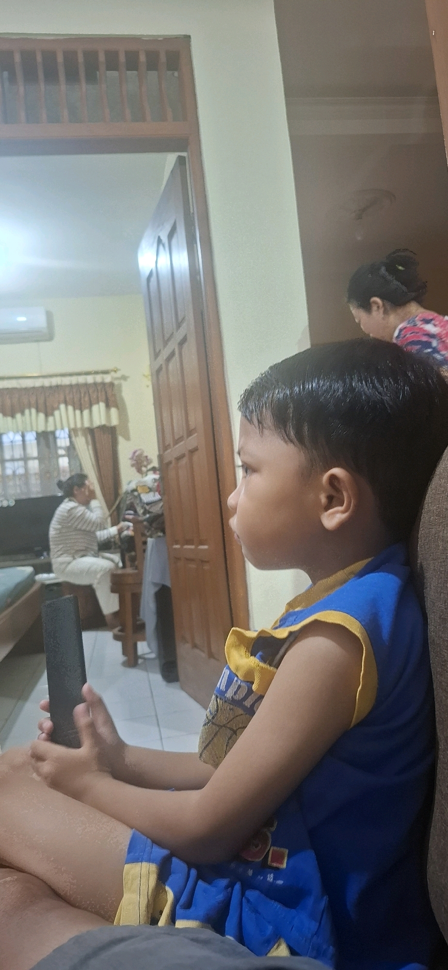 A young boy with dark hair is sitting in a living room, holding a black remote control. He is wearing a blue and yellow sleeveless shirt and blue shorts with yellow trim. He is looking off to the side, away from the camera. In the background, a woman with her hair in a bun is visible, wearing a colorful patterned shirt. Further back, another person is sitting on a stool, wearing a striped shirt and white pants, and appears to be looking at something on a table. The room has tiled flooring and wooden doors, and an air conditioning unit is mounted on the wall. The setting appears to be an indoor residential environment in Desa Gagaksipat, Indonesia.