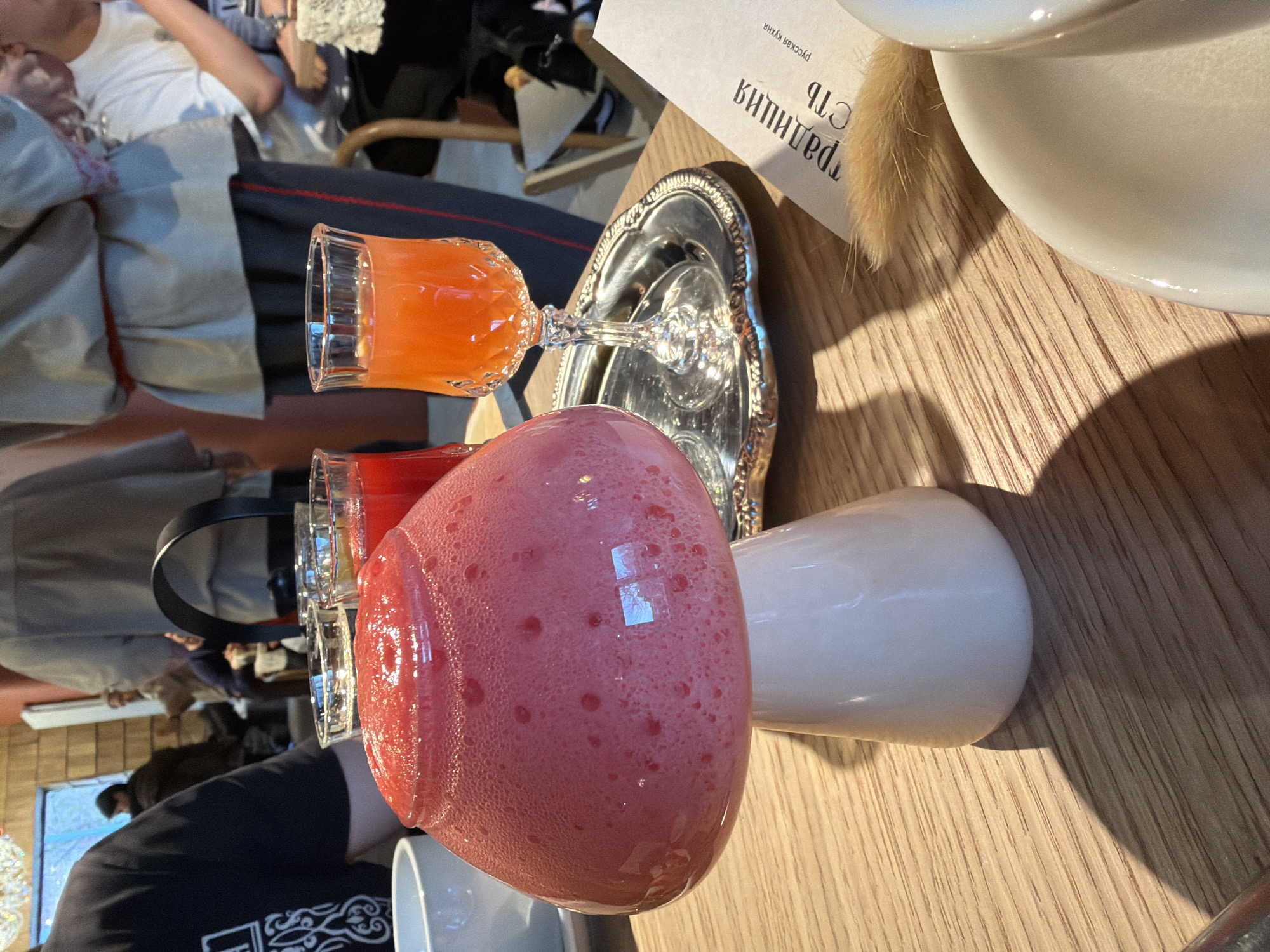 On a wooden surface, possibly a table, are several items including a pink frothy drink in a glass, an orange drink in a glass, and a silver tray. In the background, indistinct people and interior of a building can be seen. A paper with Russian text is also visible. Based on the text on the paper (русская кухня/Russian cuisine), the location is likely a restaurant in Khimki, Russia.