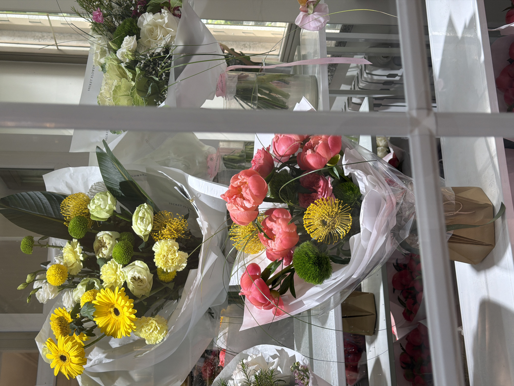 The image shows a display of bouquets of flowers behind a white-framed window. The flowers include a variety of types, such as yellow gerberas, white roses, pink peonies, and various green accents. They are wrapped in white paper and some are secured with pink ribbon. The display is possibly inside a flower shop. The location is possibly in Bishkek City, Kyrgyzstan, but this cannot be definitively determined from the image.