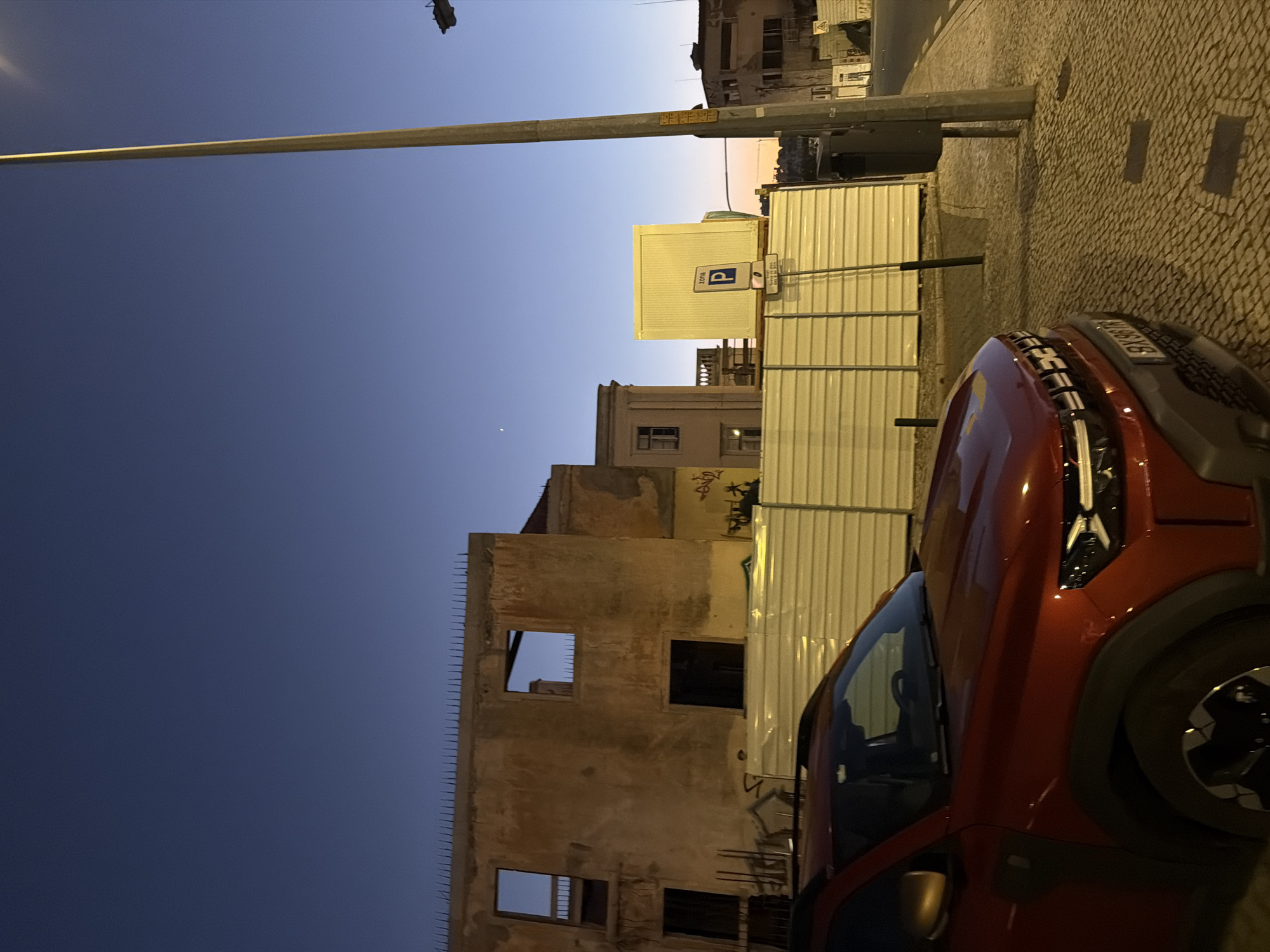 The image depicts an outdoor scene in Cascais, Portugal, during dusk. In the foreground, a red Dacia Duster car is parked on a cobblestone street. Behind the car, a temporary metal fence encloses a construction site or an area undergoing renovation. Beyond the fence, a partially demolished building with visible structural elements and empty window frames stands under a clear, deep blue sky. Further in the background, other buildings are visible, including a pale yellow portable structure with a parking sign. A tall lamppost stands prominently to the right of the image. The overall atmosphere suggests a transitional period between day and night, with artificial lights starting to illuminate the urban landscape. There are no people visible in the frame.