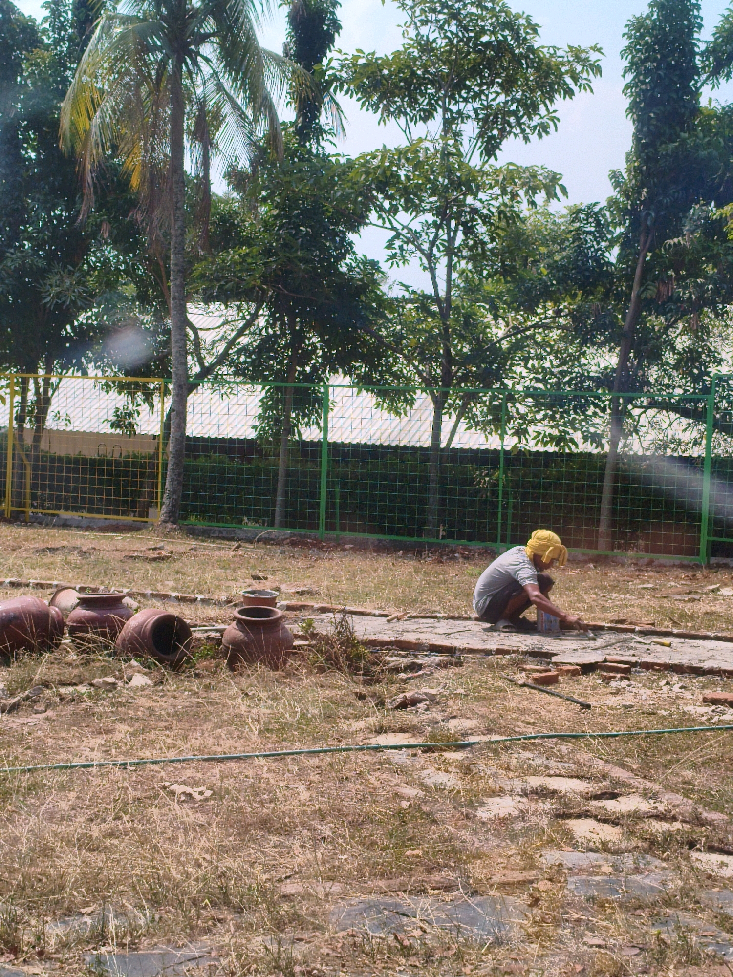 In Sukaraja, Indonesia, a person wearing a yellow head covering and a grey shirt is crouched on a paved area, appearing to be working with cement or mortar. Several earthenware pots are arranged on the dry, grassy ground to the left of the person. Behind them, a wire mesh fence, partially yellow and partially green, separates the area from a row of trees and structures. The sky is bright and clear.