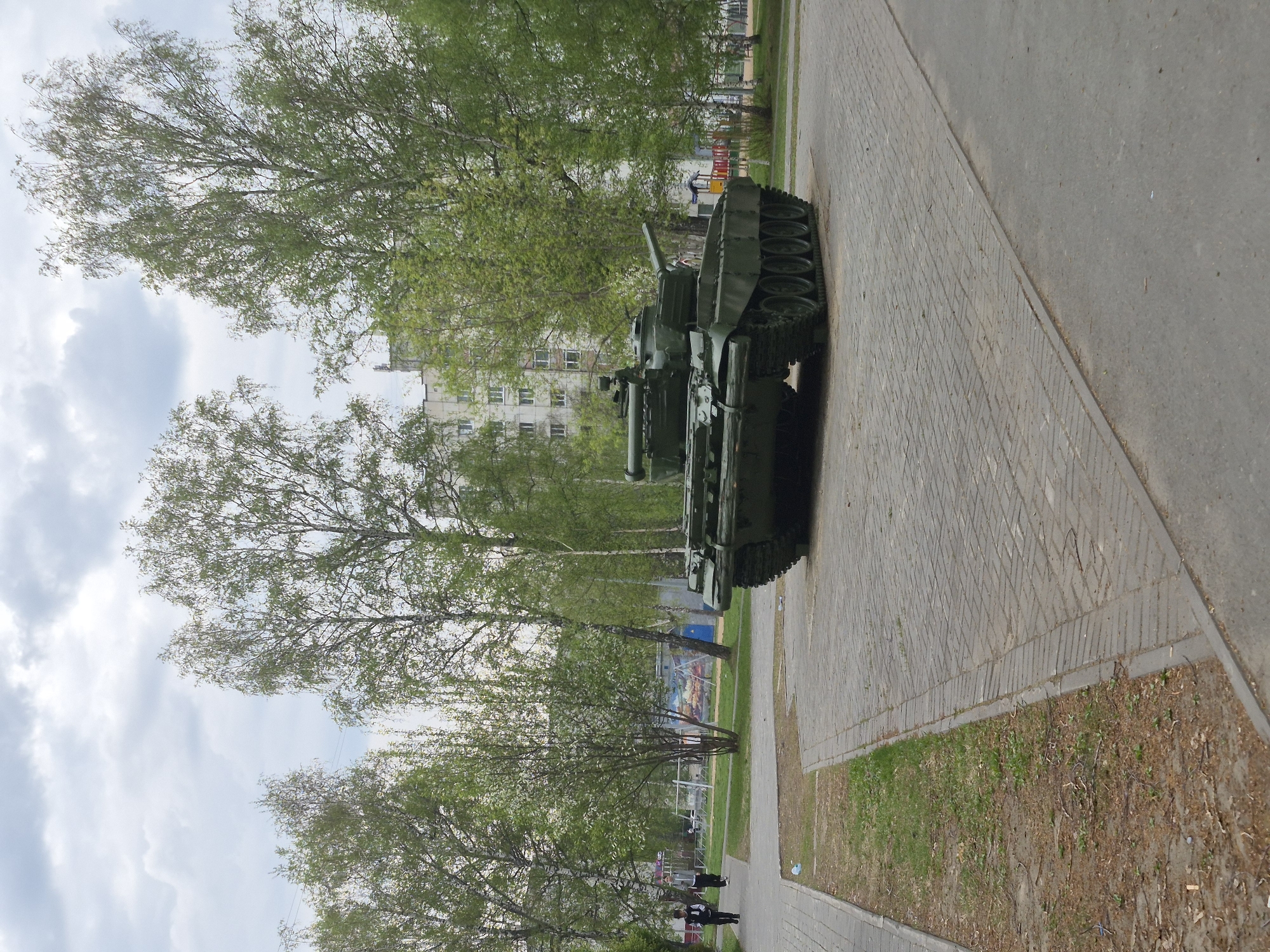 A slightly angled, long shot shows two green battle tanks parked on a paved surface, likely a street or walkway, in Tobolsk, Russia. The tanks are the main focus, positioned side-by-side, with their turrets visible. Behind the tanks, trees with light green foliage line the area. In the background, a light-colored building stands, and a sports field with fencing can be seen. At the bottom of the frame, two individuals are walking away from the camera on the sidewalk, near more trees. The sky is visible through the trees, with a mix of blue and white clouds. The image appears to be taken during the daytime in spring or early summer.