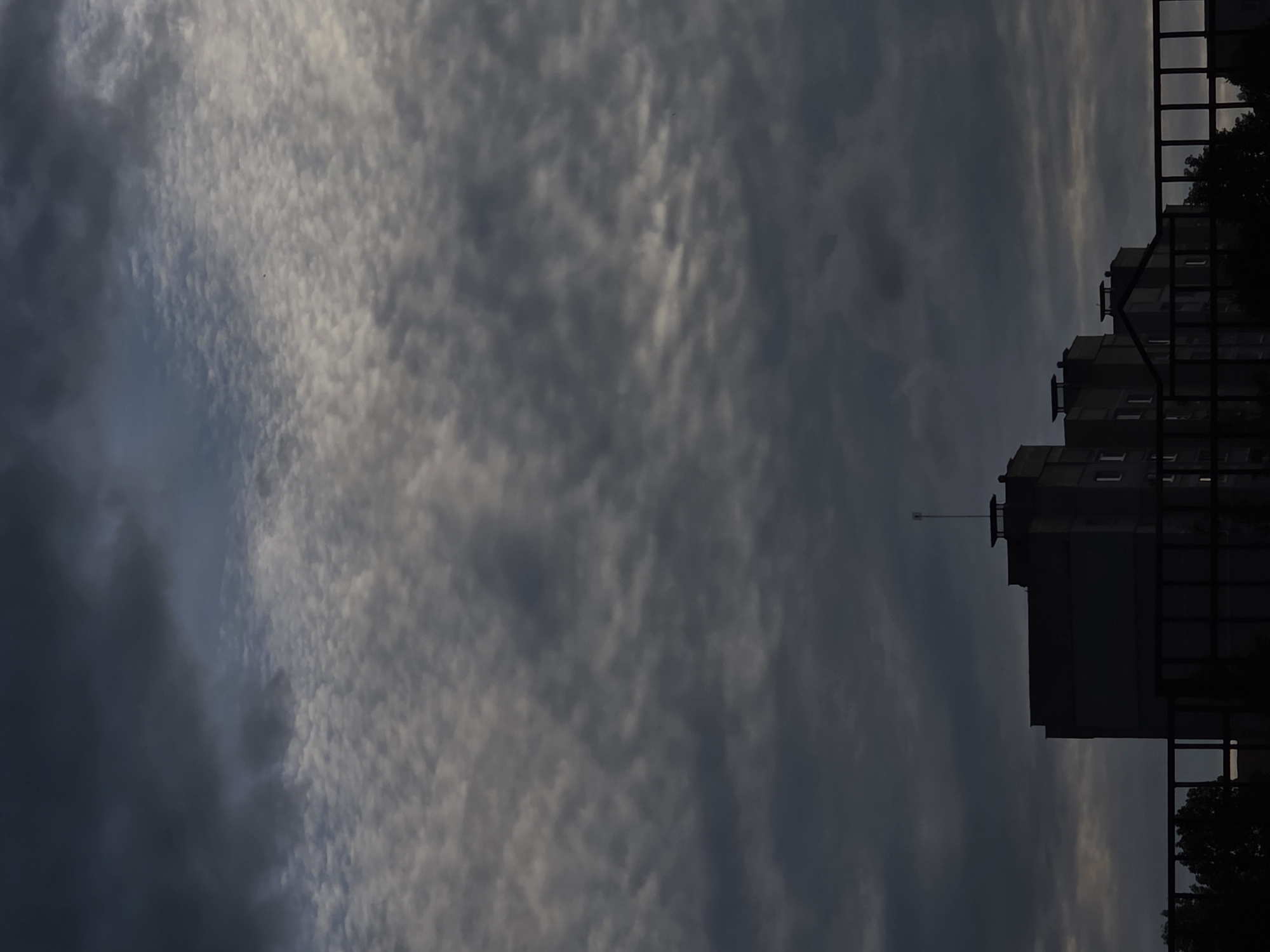 An exterior, eye-level photograph captures a somber urban landscape under an overcast sky. The upper two-thirds of the image are dominated by a dense cloud cover, consisting of dark grey, heavy clouds in the upper left transitioning to lighter, textured stratocumulus or altocumulus clouds with scattered white-grey patches across the mid-sky. The lower third of the image features a series of multi-story buildings, largely silhouetted against the ambient light. The most prominent building on the left exhibits a tiered, flat-roof design with a thin, vertical antenna-like structure on its highest point. Adjacent buildings to the right share a similar architectural style. Several windows are visible on the building facades, with a few emitting faint light from within. A dark metal railing or fence traverses the immediate foreground, obscuring the lower portions of the buildings. Dark, indistinct foliage, likely trees, is visible behind the fence and in front of the building line. No individuals are present. The scene is set in the city of Unknown, Unknown, under conditions suggesting dusk or a heavily overcast day.