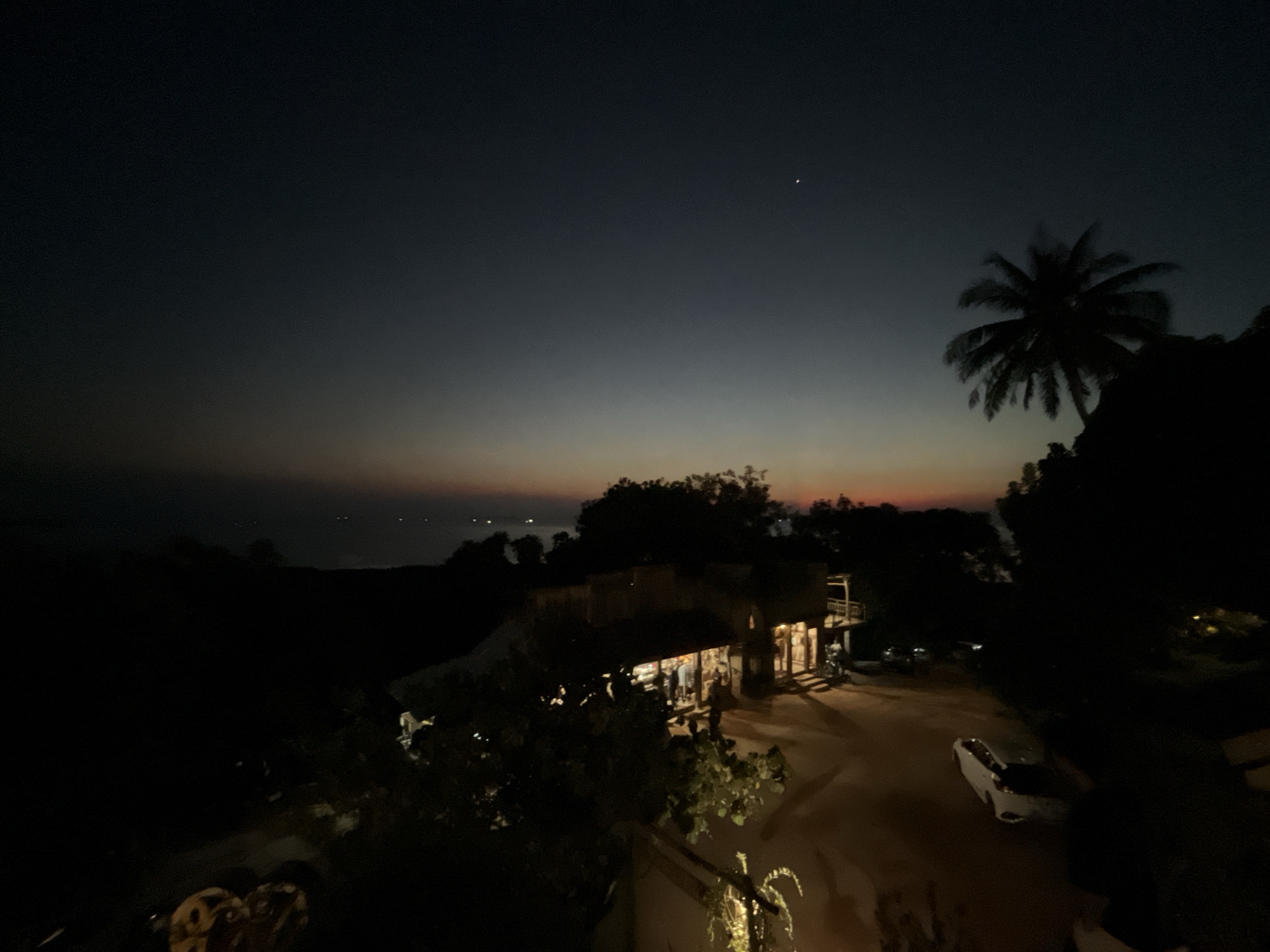 This is a nighttime aerial view of a coastal area in Karimunjawa, Indonesia, just after sunset. The sky transitions from a deep twilight blue at the top to faint hues of orange and pink near the horizon, suggesting the last remnants of daylight. A single bright star is visible in the upper right quadrant of the sky. In the foreground, a silhouetted palm tree stands against the dusky sky.
Below, a cluster of buildings with illuminated windows indicates a small settlement or resort. Lights are on inside the structures, and there appears to be some activity, with figures visible through the windows, possibly people dining or socializing. A dirt or gravel path winds through the scene, with a white car parked on it. The overall impression is one of a tranquil evening in a tropical location. The scene evokes a sense of peace and relaxation, characteristic of a vacation spot. There is no visible text in the image.