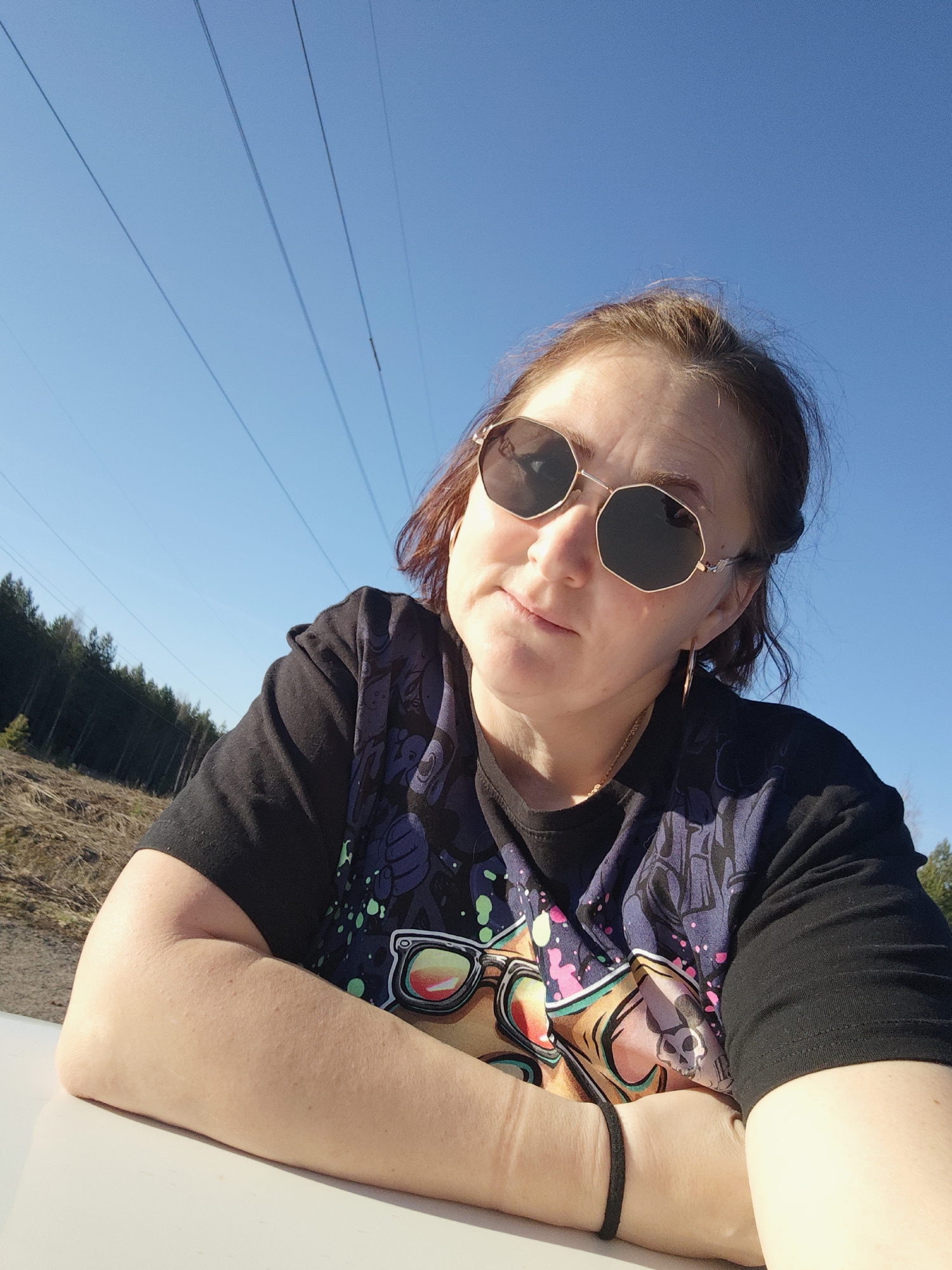 The image features a woman posing for a photograph outdoors on a bright, sunny day. She has light brown hair, pulled back, and is wearing octagonal-shaped sunglasses with gold frames. She is dressed in a black short-sleeved t-shirt adorned with colorful, cartoon-like graphics, which appear to include stylized faces or characters in vibrant hues of purple, green, pink, and yellow. A thin gold chain necklace and a black bracelet on her left wrist are also visible.
The setting is characterized by a vast, clear blue sky, stretching above. Multiple thin power lines traverse the sky, suggesting the presence of electrical infrastructure. In the background, a dense line of coniferous trees forms a forest, bordering an area of sparse, dry ground. The woman is positioned close to the camera, resting her forearms on a light-colored surface, possibly the hood or roof of a vehicle, or a low wall.
She is looking directly or slightly past the camera, with a neutral expression; her eyes are hidden by the sunglasses, but a slight furrow in her brow is visible. The strong sunlight casts distinct shadows, indicating it is daytime. The overall scene suggests a casual outdoor portrait taken in a natural or semi-rural environment, consistent with locations in Chaikovsky, Russia. No discernible text is visible on her clothing or in the background.