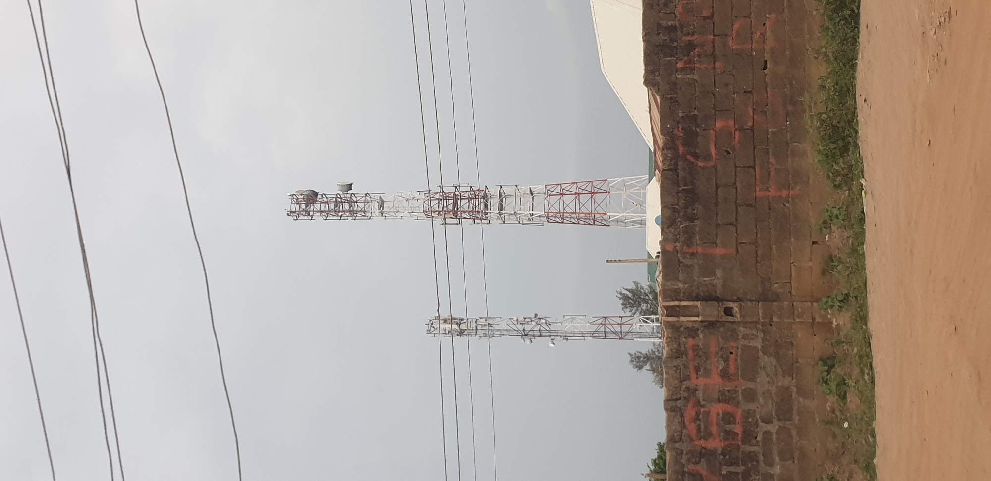 In Sabon Tasha, Nigeria, two tall telecommunication towers stand prominently against a cloudy sky. The tower on the right is red and white, while the one on the left is primarily white with red accents. Both are lattice structures with antennas and equipment mounted at various levels. In the foreground, a low brick wall is visible, marked with large orange symbols or numbers. Sparse green vegetation grows along the base of the wall. A dirt road or path lies in front of the wall. The overall scene suggests an urban or semi-urban environment focused on communication infrastructure.