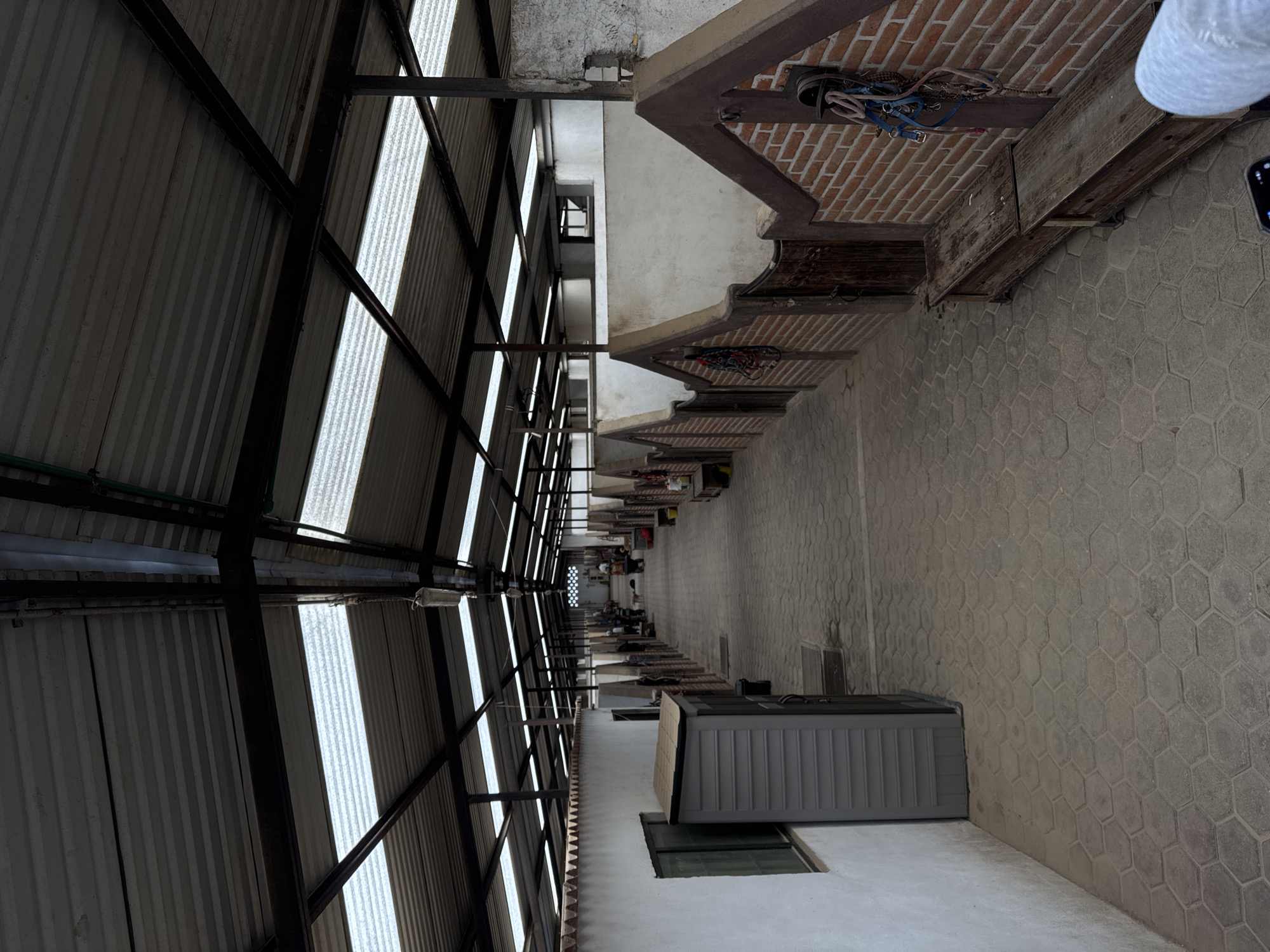The image depicts a long, narrow alleyway in San Gregorio Atzompa, Mexico. The alley is bordered on one side by a tall corrugated metal structure with support beams, likely forming a roof or wall. The other side features a series of overhanging ledges or eaves constructed with brick and wood, suggesting adjacent buildings. The ground is paved with hexagonally patterned blocks. A large gray plastic storage container sits against a white wall with a visible window frame near the bottom of the image. In the distance, several people are visible standing or walking near the end of the alley. Overhead, various items such as a horse harness and other objects are stored under the overhanging eaves.