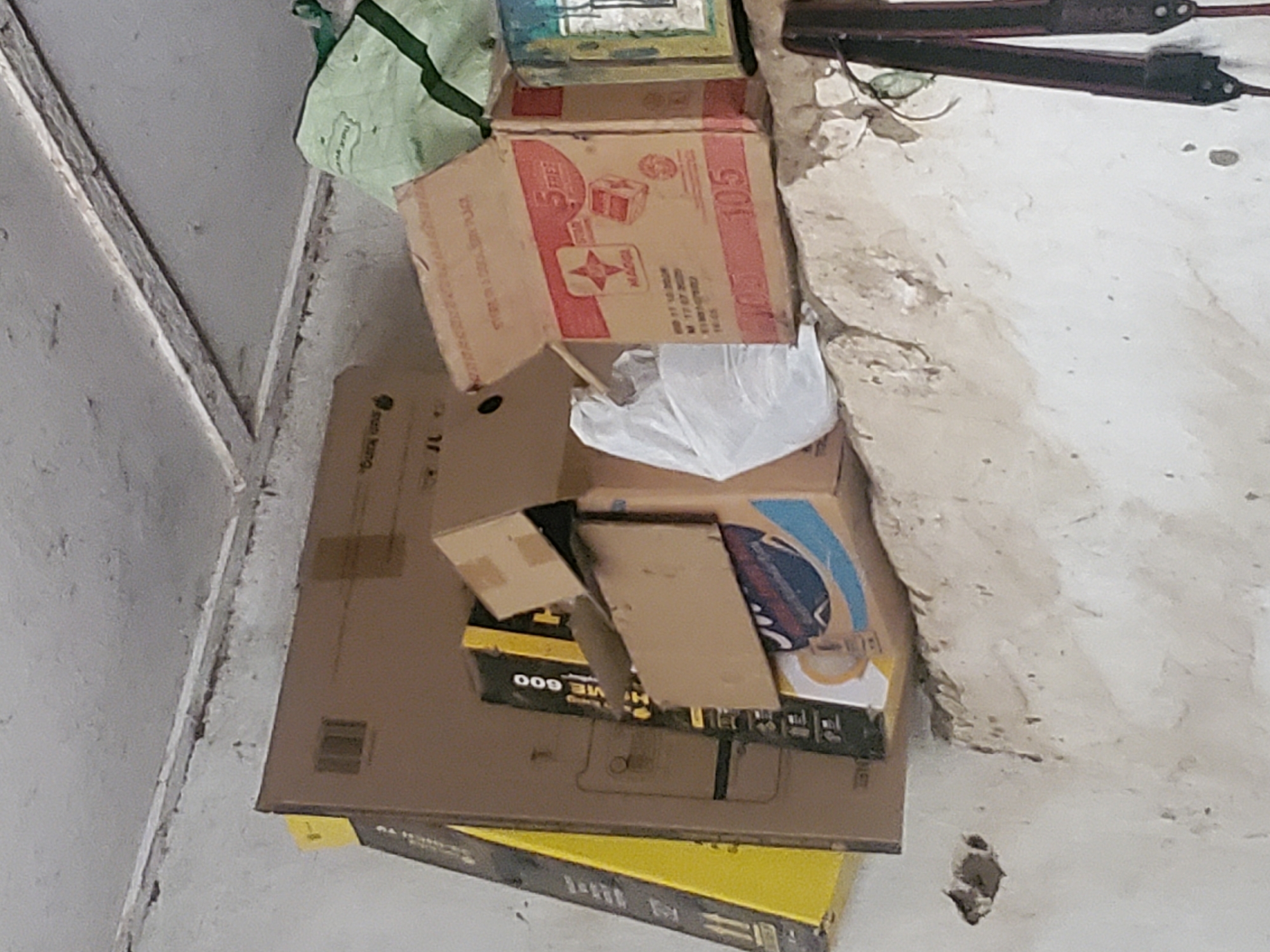The image depicts a cluttered storage area. Several cardboard boxes of various sizes are stacked on a rough, light-colored ledge. One prominent box is red and white, featuring the brand "MAGGI" and the number "105". Another box is partially visible to the left, with yellow and black markings. A white plastic bag is also present among the items. In the background, a green fabric item is partially visible. The wall appears to be made of concrete or plaster, with some signs of wear and tear, including holes and chipped paint. The ceiling is a textured, light gray surface with visible beams or structural elements. The overall impression is that of an unorganized storage space, possibly a garage or a storeroom. This scene is located in the city of Kabwir, Nigeria.
