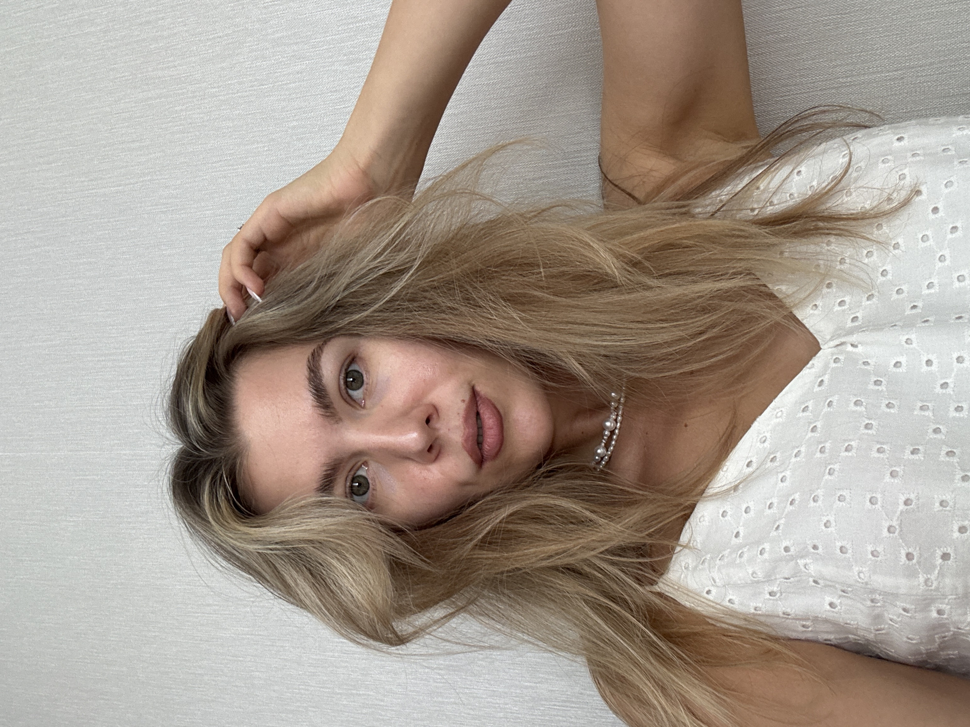A young woman with blonde hair pulled back from her face stands in front of a textured, off-white wall. She is wearing a white eyelet camisole and a pearl necklace. Her left hand is raised to her head, and her right arm is bent behind her. The lighting suggests an indoor environment.
