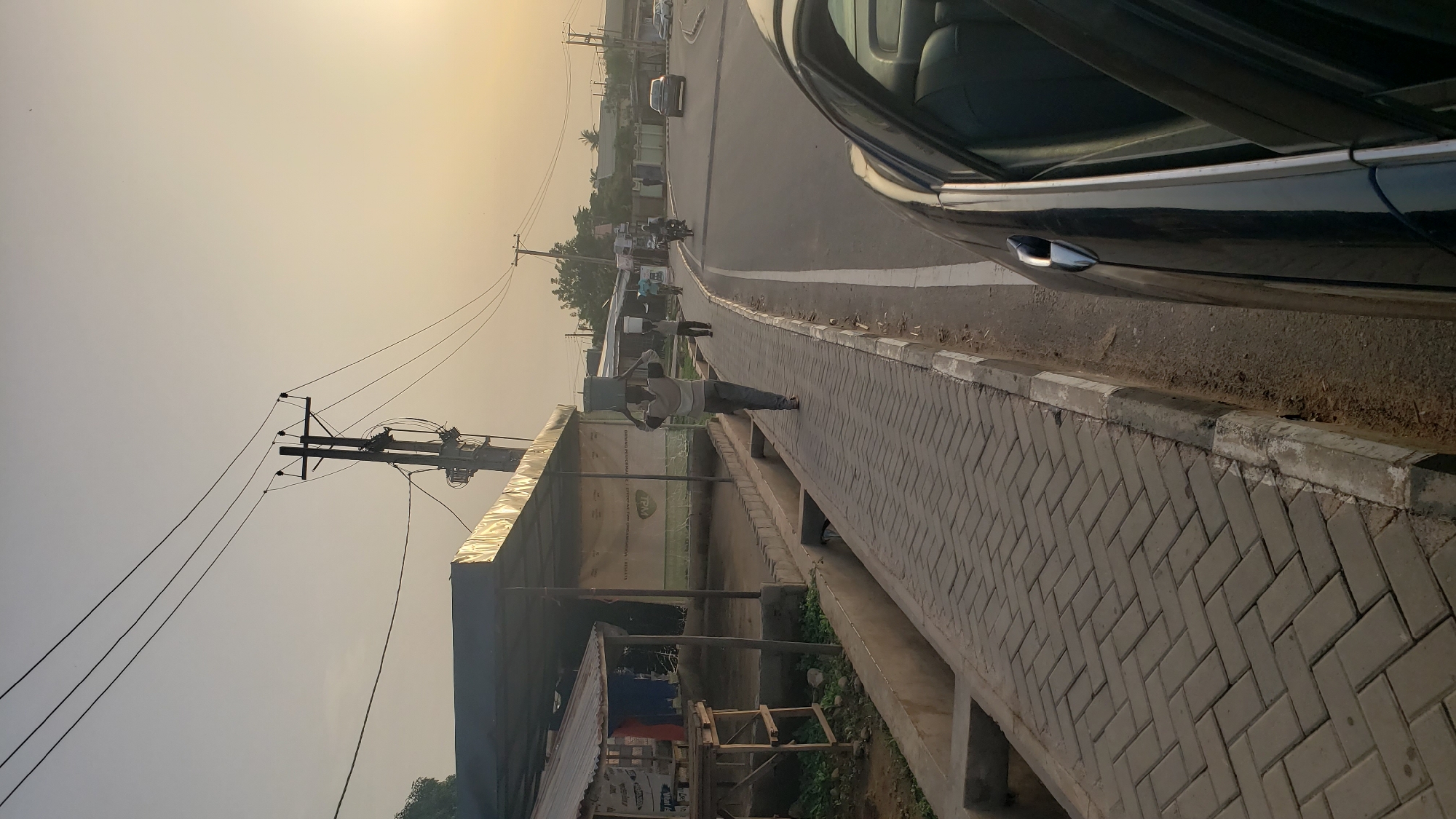 The image depicts a street scene in Narayi, Nigeria. A man is walking along a paved walkway next to a road. A black car is parked on the side of the road, partially visible in the foreground. Power lines run alongside the street. A small, covered structure stands next to the walkway, with various objects underneath. Other people and vehicles are visible in the distance on the road. The sky is hazy and bright, suggesting either sunrise or sunset.