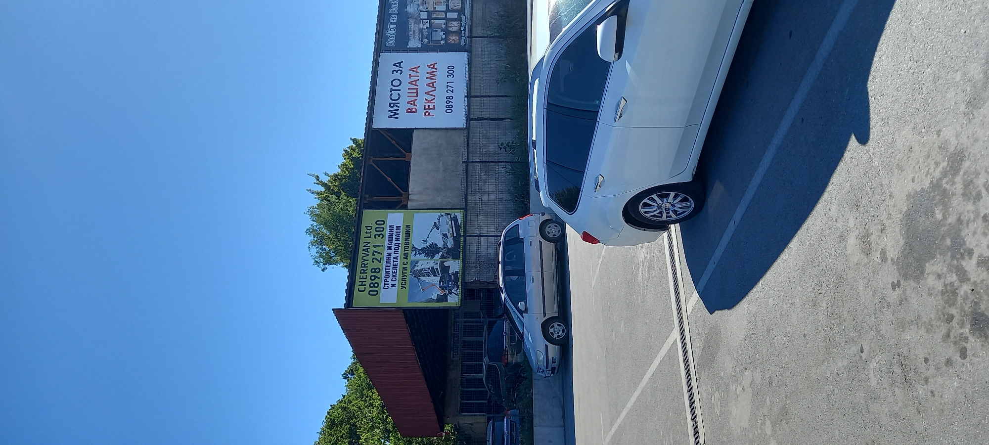 The image shows a view of a parking lot in front of a building with advertisements on its exterior wall. The sky is clear and blue. In the foreground, the rear of a white station wagon is visible, parked in a designated spot. To the left, a silver station wagon is parked in another spot, and further left, other vehicles are partially visible.
The building behind the parking lot appears to be a commercial or industrial structure with a dark-colored roof and a reddish-brown corrugated metal wall on the left side. Several large advertisement posters are attached to the front of the building.
One prominent green poster displays the text "CHERRYVAN Ltd. 0898 271 300" and below it, "СТРОИТЕЛНИ МАШИНИ И СКЕЛЕТА ПОД НАЕМ УСЛУГИ С АВТОБИШКИ", which translates to "BUILDING MACHINERY AND SCAFFOLDING FOR RENT SERVICES WITH AUTOS". This poster also features illustrations of construction equipment and a building.
To the right of the green poster, a white poster reads "МЯСТО ЗА ВАШАТА РЕКЛАМА 0898 271 300", meaning "SPACE FOR YOUR ADVERTISING".
Further to the right, another poster is partially visible and appears to be advertising baked goods or food products.
The context of the location being the city of Silistra, Bulgaria, suggests that this is a commercial area possibly related to equipment rental and advertising services within the city. The overall scene depicts a typical day in an urban environment with commercial activity.